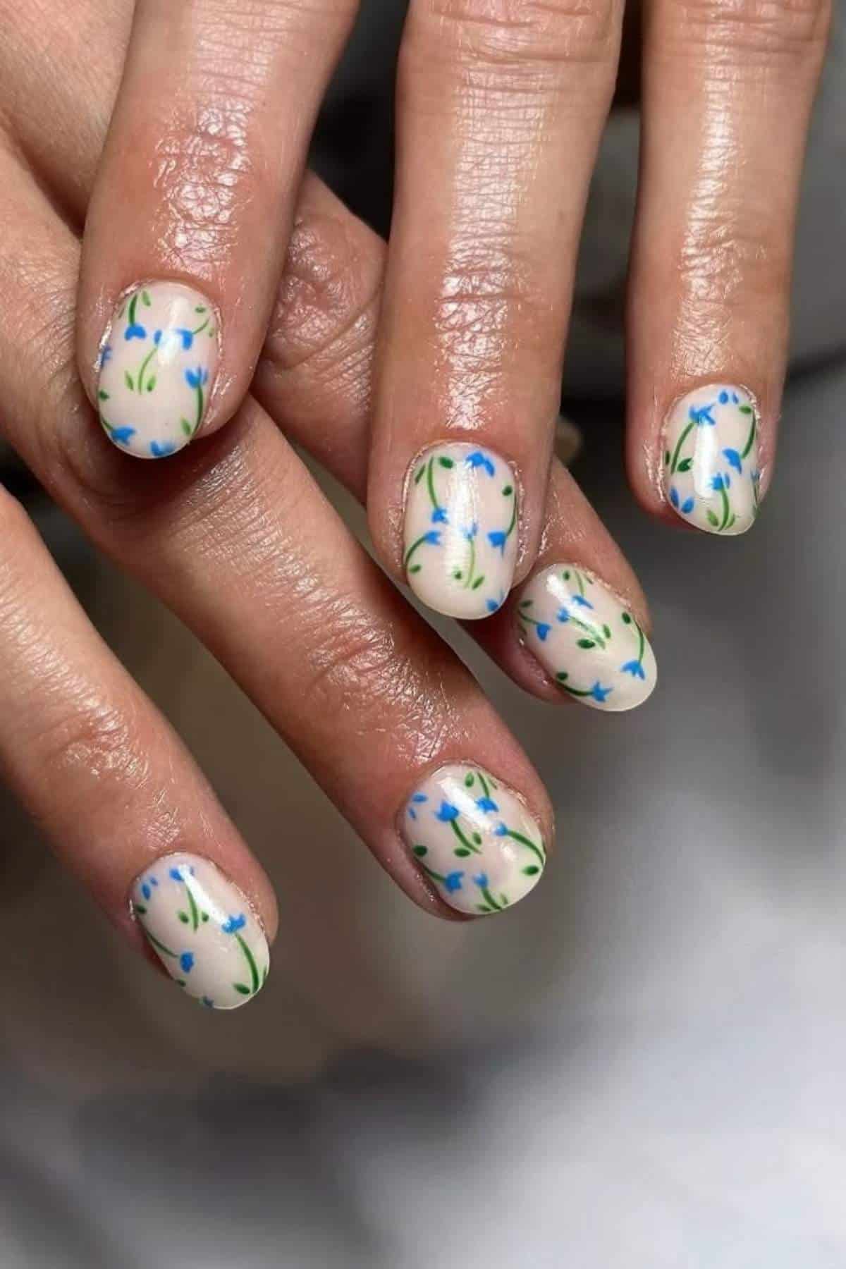 A close-up of hands showing fingernails painted with a light base color and decorated with small blue flowers and green leaves.