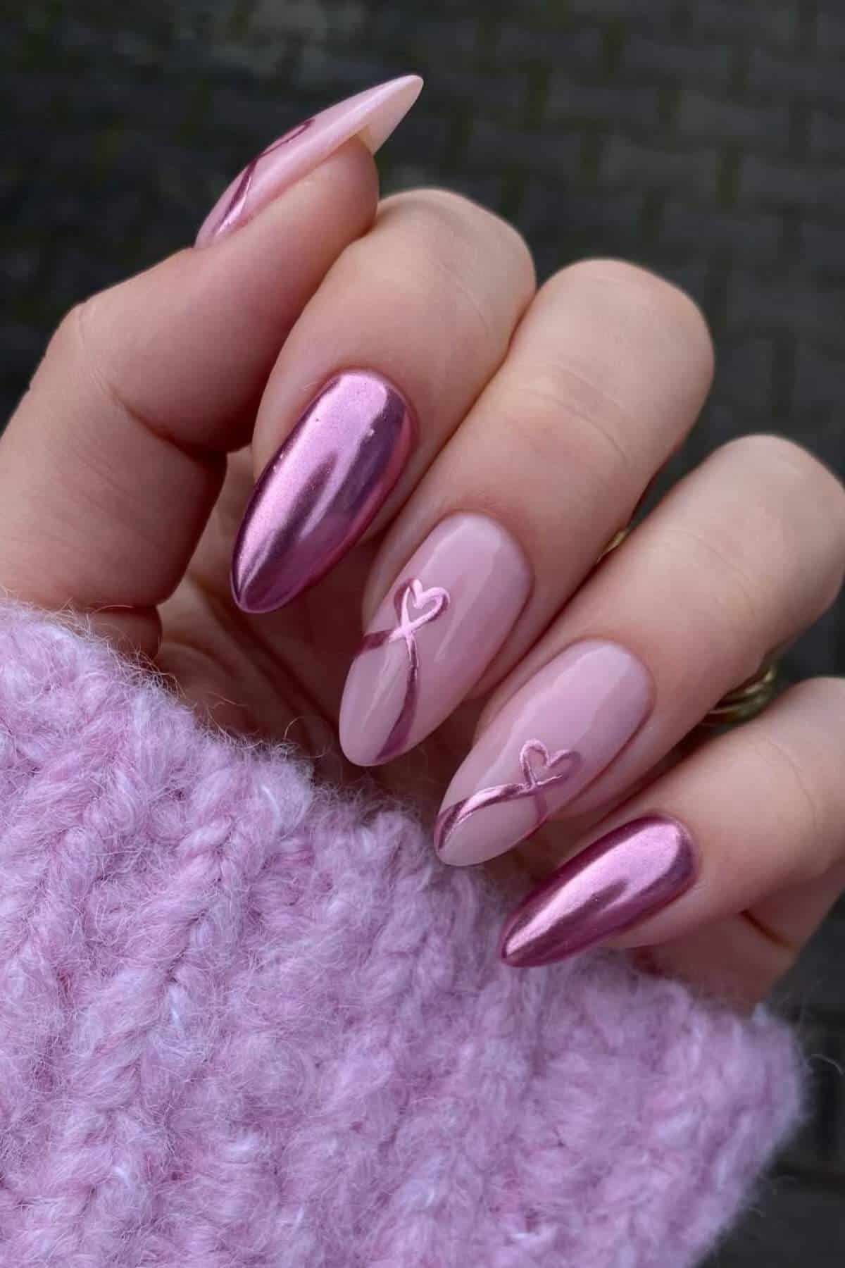 A hand with almond-shaped pink nails featuring metallic and glossy finishes, heart designs, and a light pink knitted sweater sleeve.