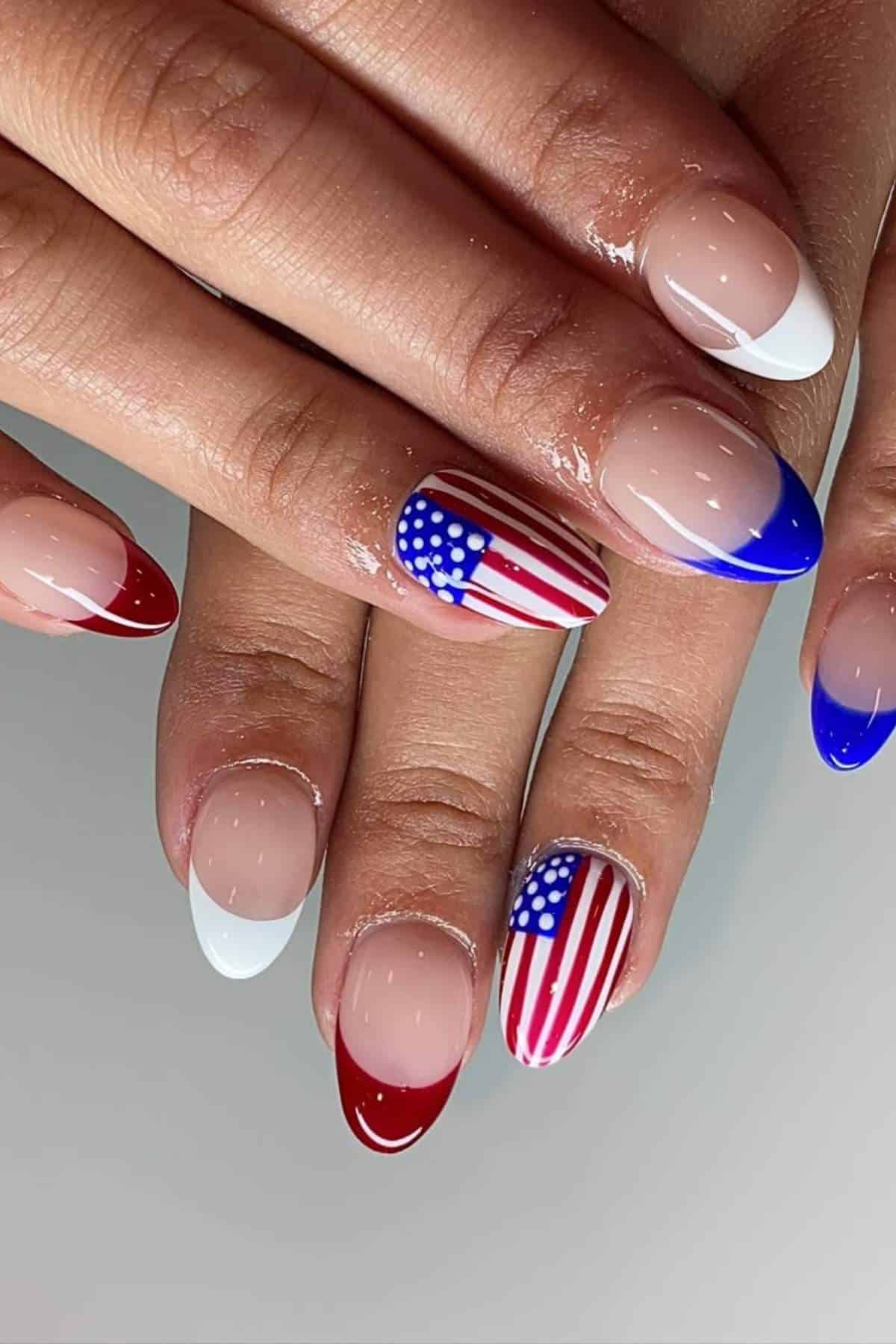 Hands with almond-shaped nails featuring French tips in red, white, and blue, with two nails painted with the American flag design.
