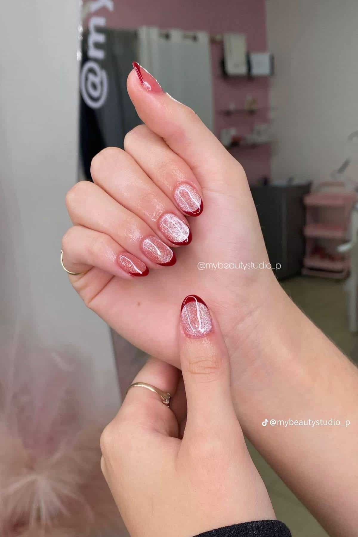 A hand with almond-shaped nails featuring a red French tip and glittery pink designs, shown in a nail salon setting.