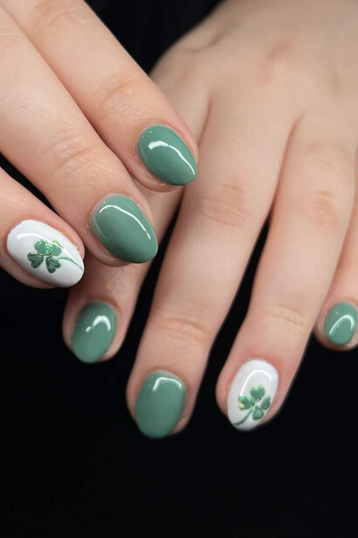 Hands with short, oval-shaped nails painted green and white; accent nails feature small green shamrock designs on a white background.