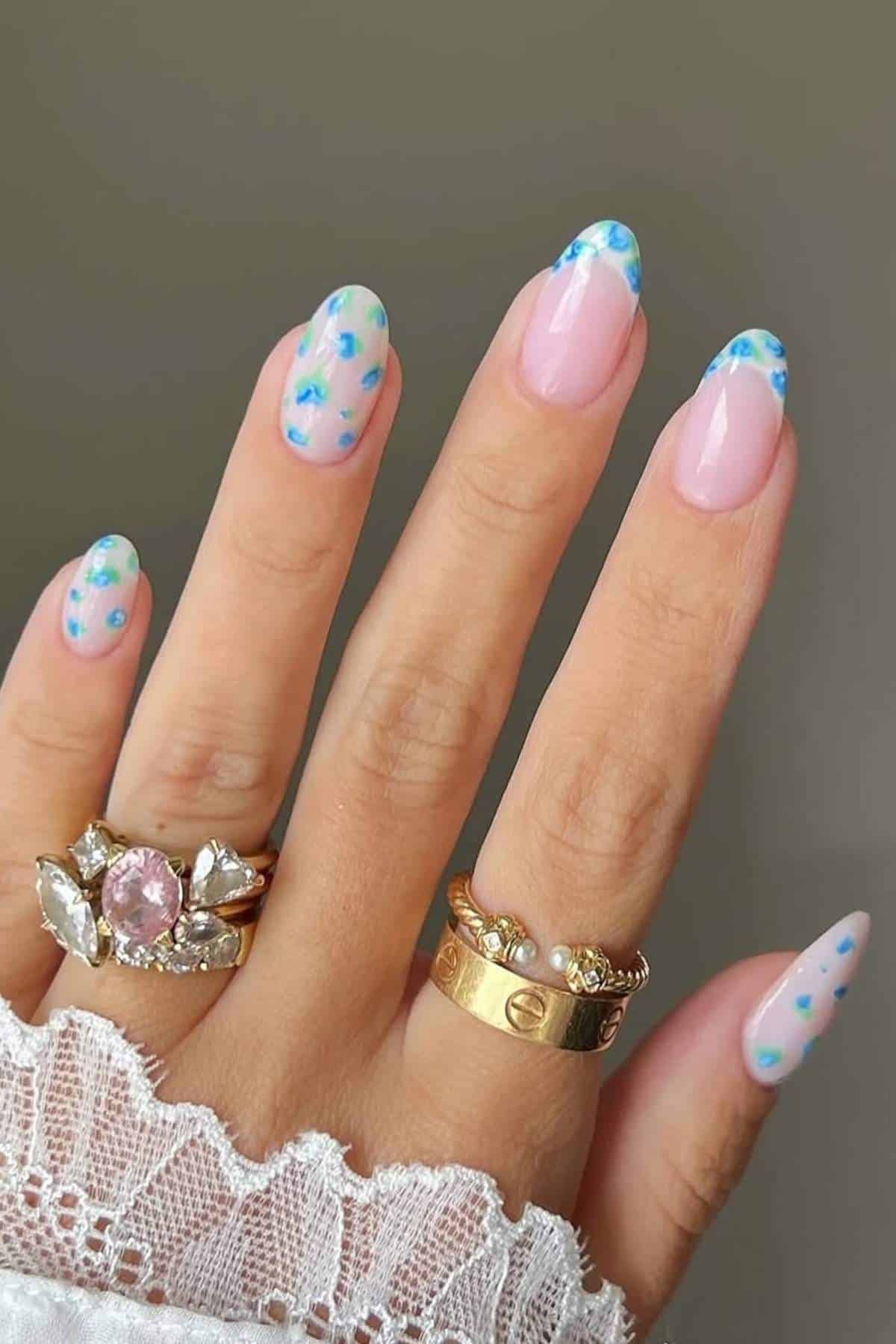 A hand with long, almond-shaped nails featuring a pink base and blue-green floral tips. The person wears gold rings with gemstones and a white lace sleeve is visible.
