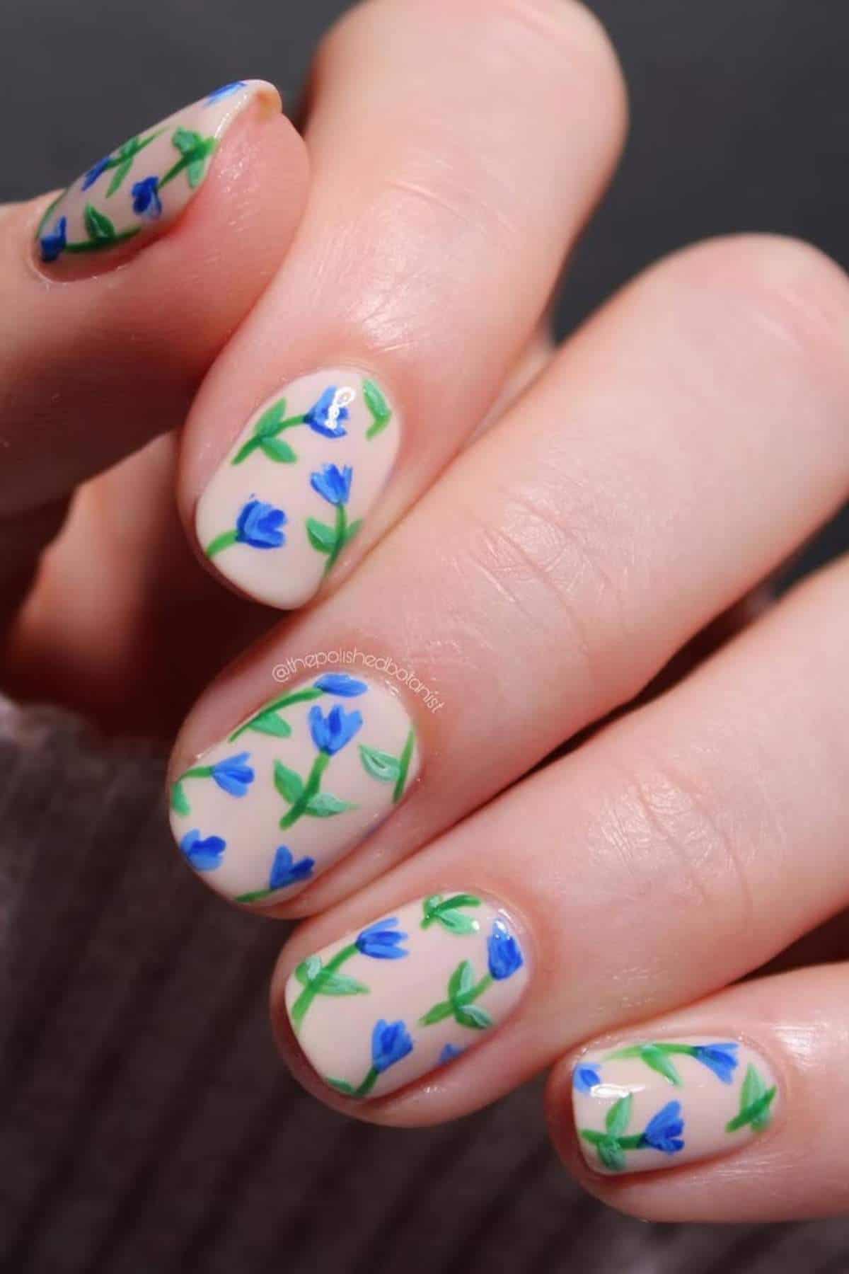 A hand with short nails painted with a clear base and decorated with small blue flowers and green leaves in a floral nail art design.