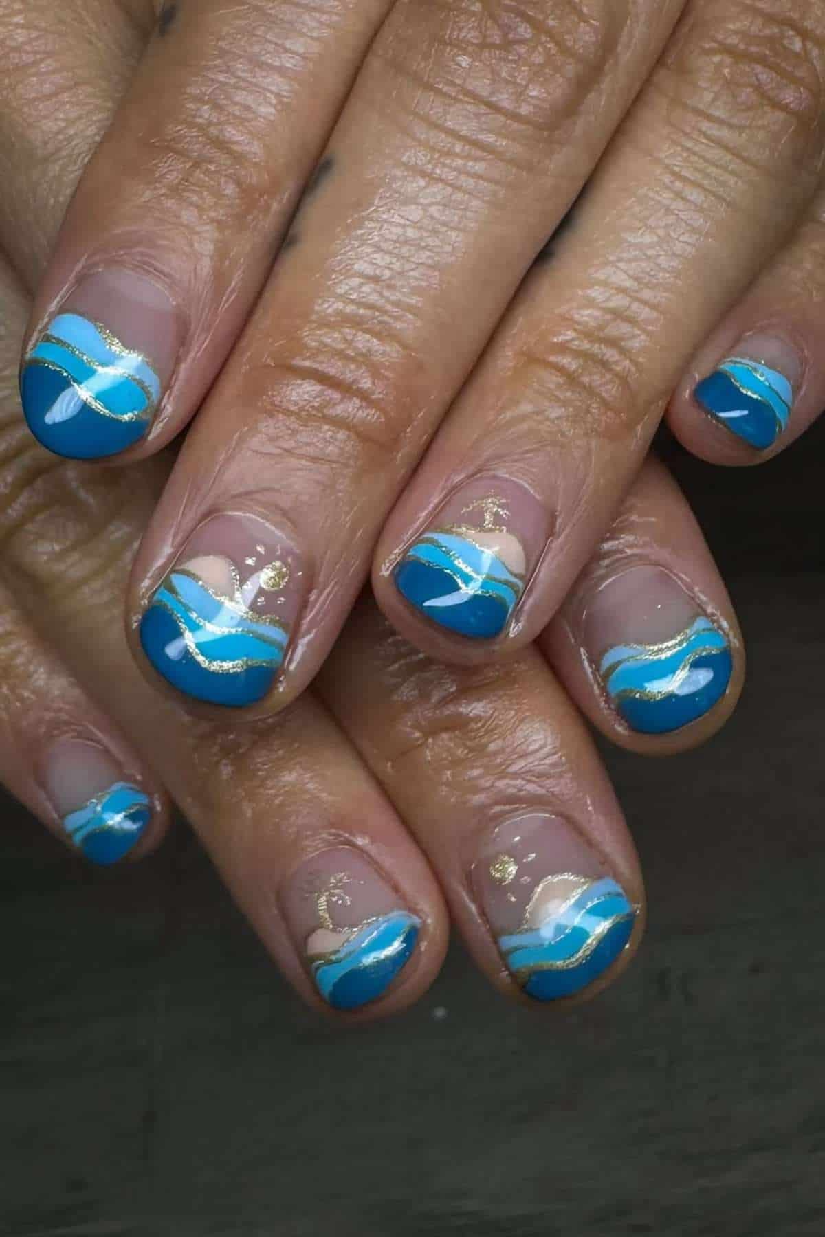 Close-up of fingernails with blue, white, and gold wave nail art on natural nails, featuring a mix of curved lines and accents.