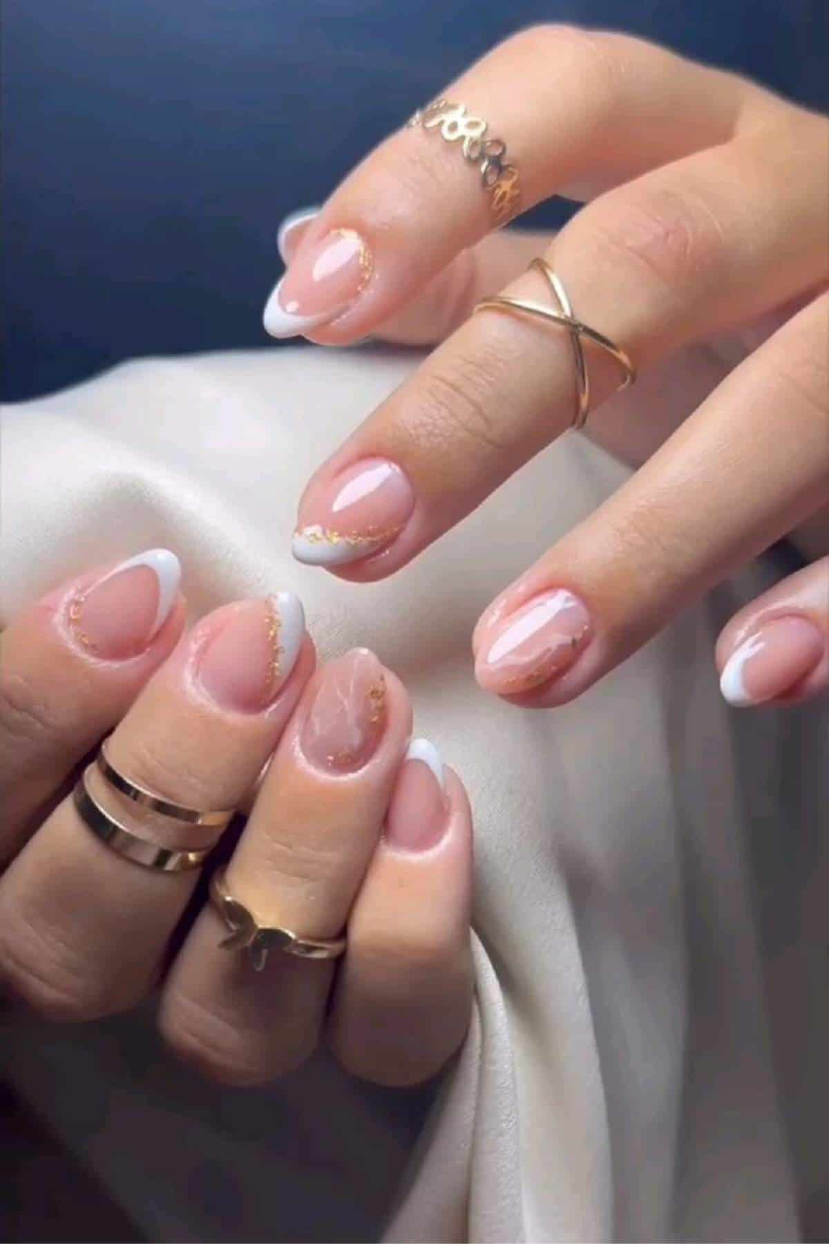 A hand with almond-shaped nails featuring a pink base, white French tips, gold accents, and several gold rings, holding a white fabric.