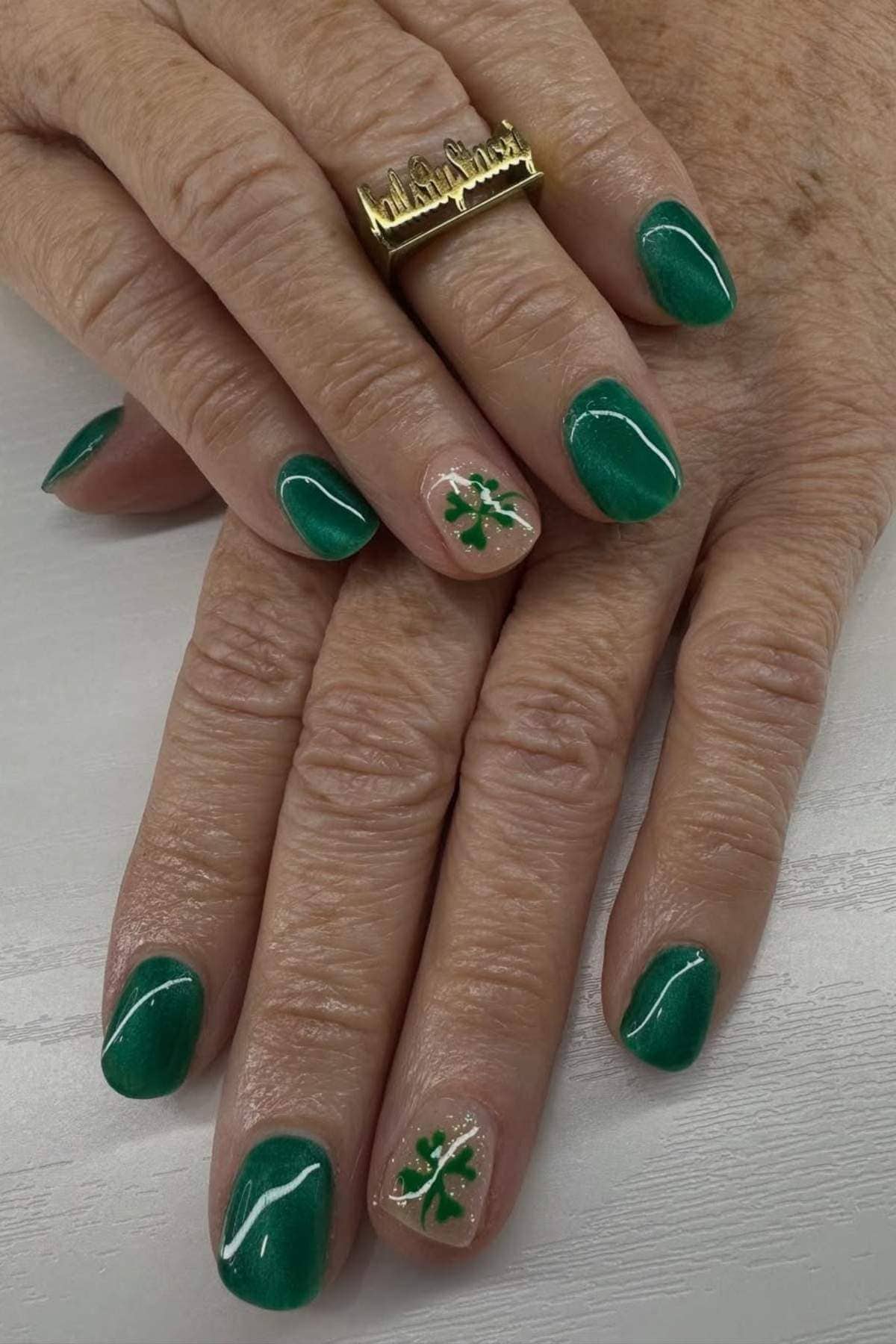 Hands with green-painted nails; ring fingernails feature a shamrock design on a glittery nude base. One hand wears a gold cursive ring.