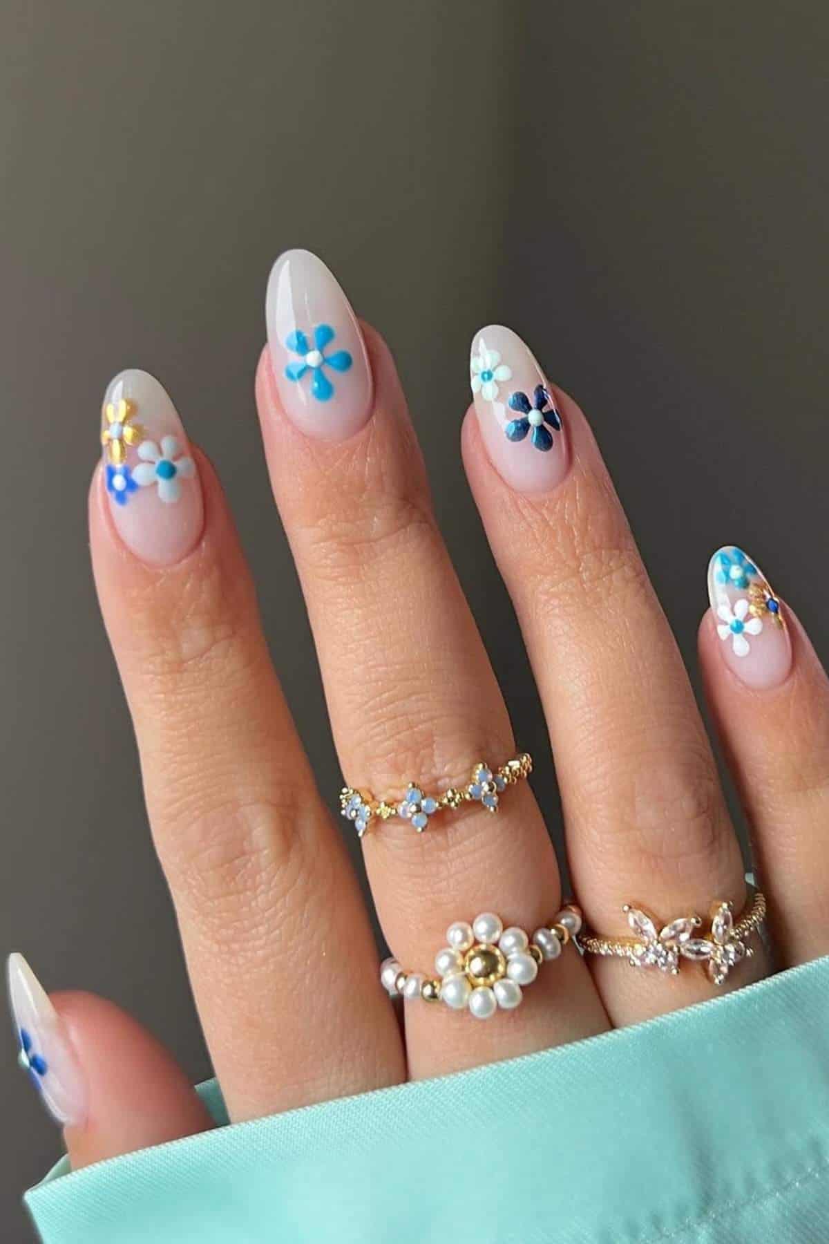A hand with long, almond-shaped nails painted with small blue and yellow flower designs, wearing several gold rings with pearls and gemstones.