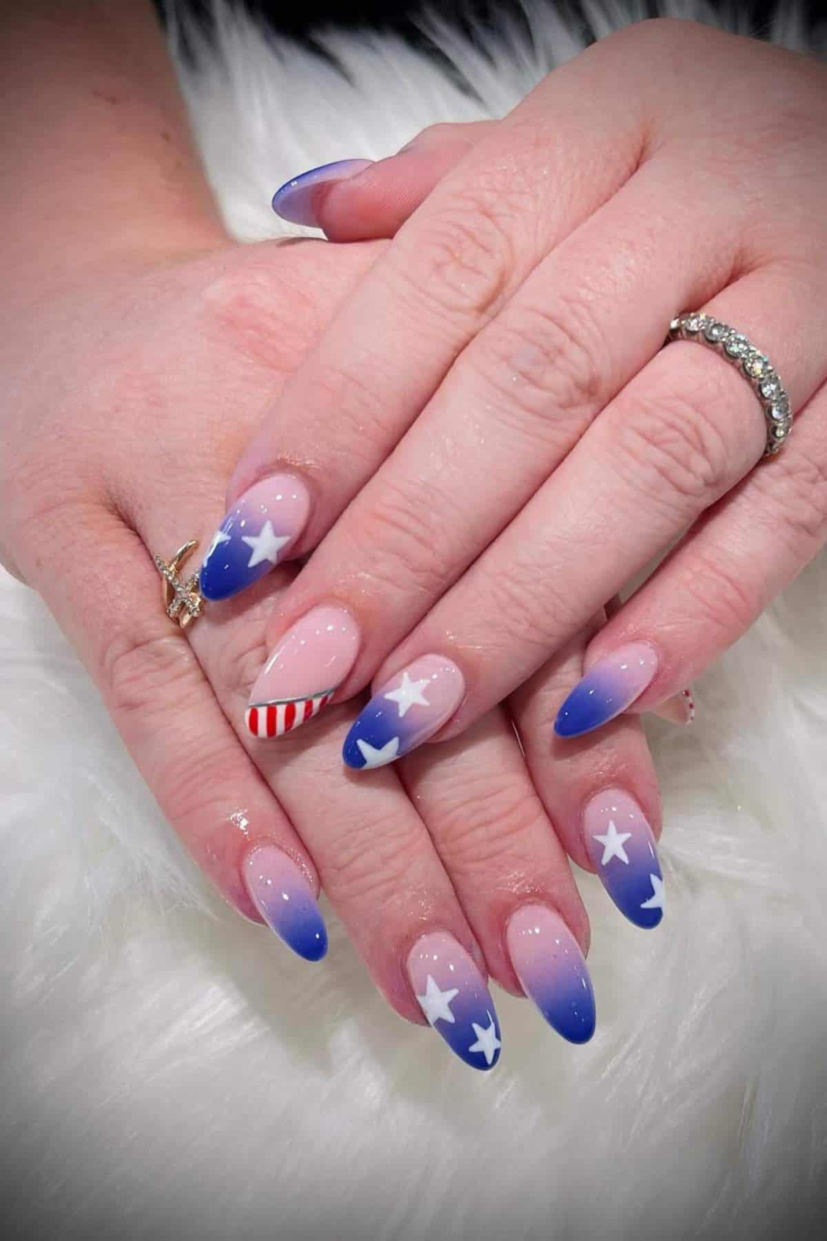Hands with almond-shaped nails featuring a patriotic design: blue tips, white stars, and one nail with red and white stripes, resting on a white surface.