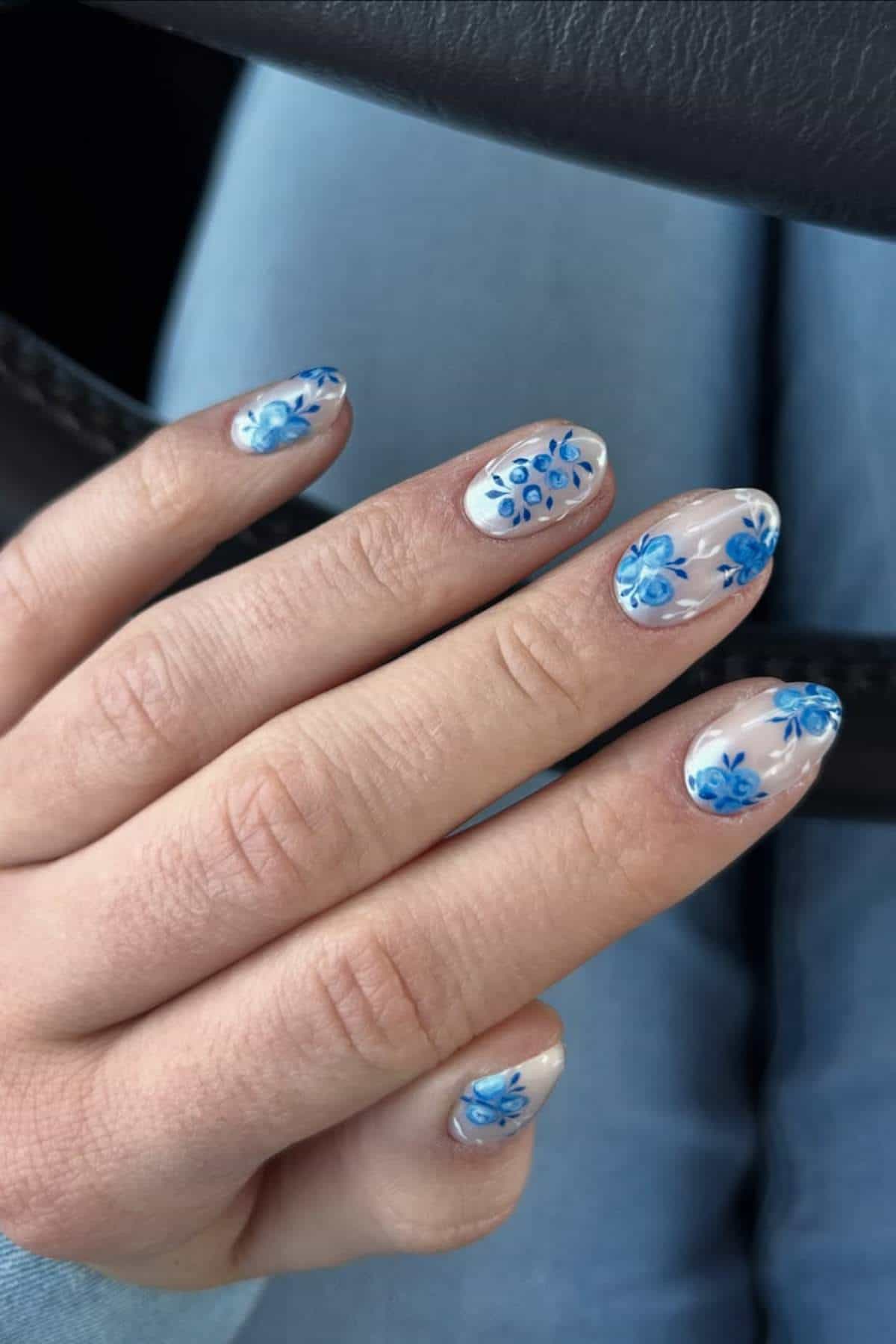 A hand with almond-shaped nails featuring clear polish and blue floral designs, resting on a light blue denim-clad leg.