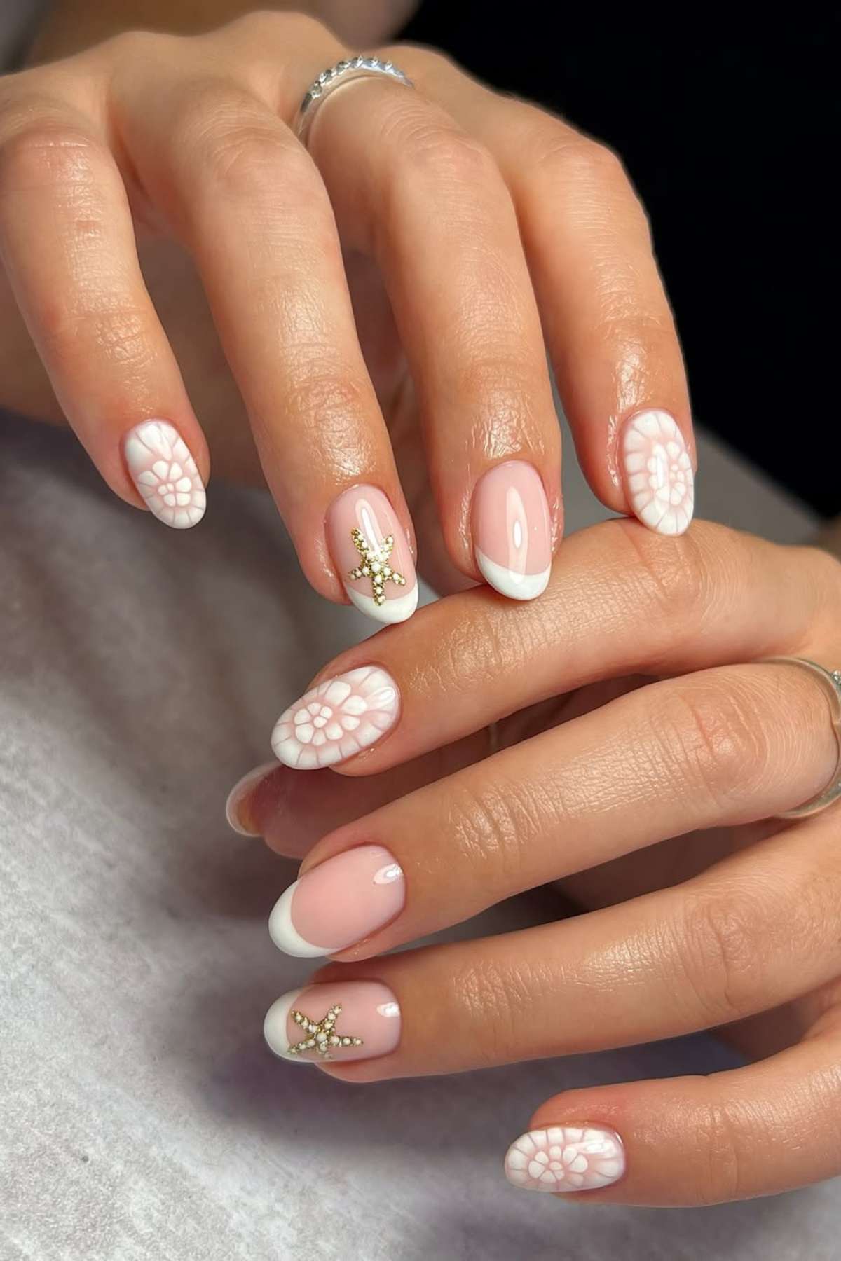 Hands with manicured nails featuring pink and white French tips, floral lace designs, and small gold starfish embellishments on some nails.