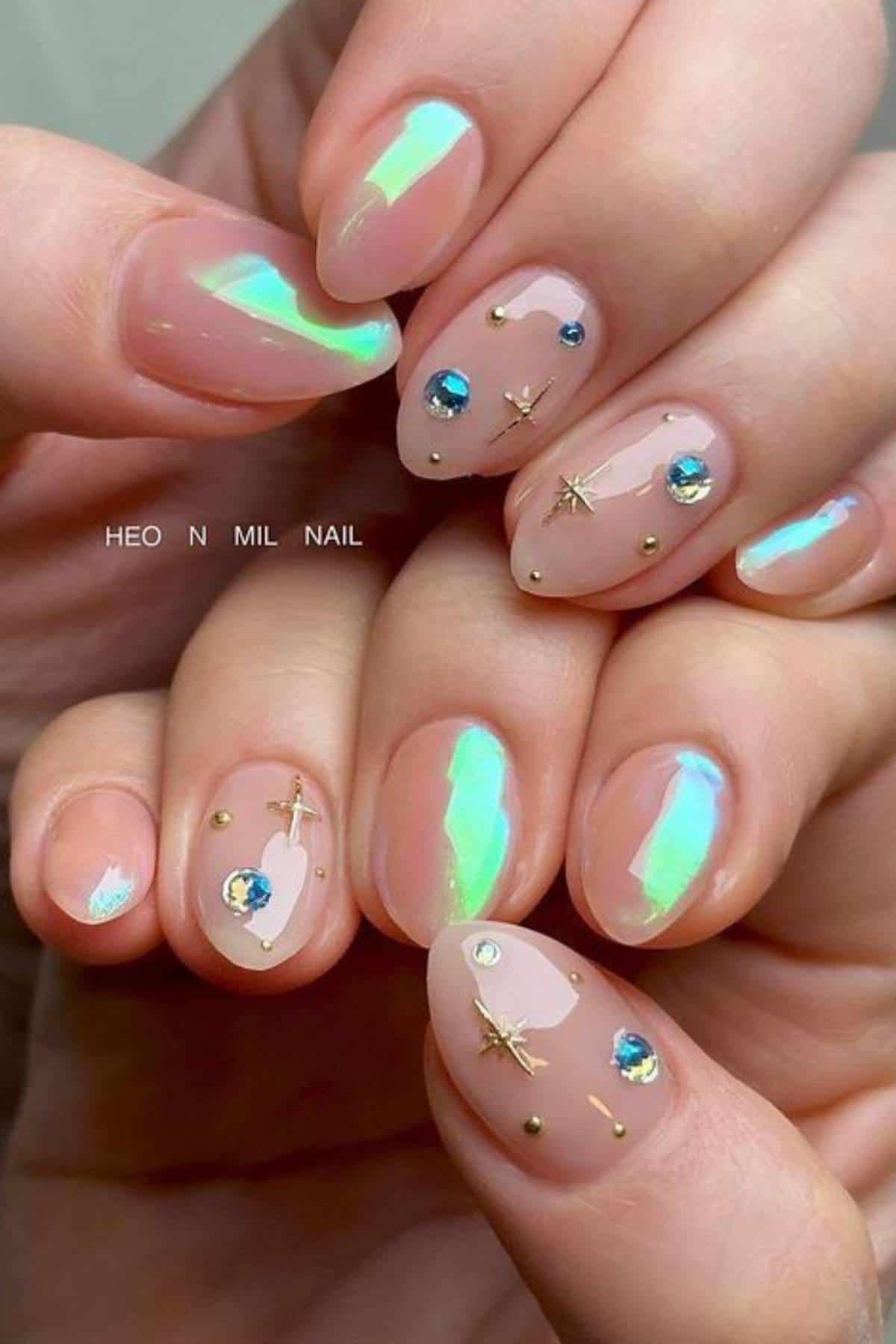 Hands with almond-shaped nails featuring a glossy iridescent finish, blue gem accents, and small gold star and dot decorations on a nude base.
