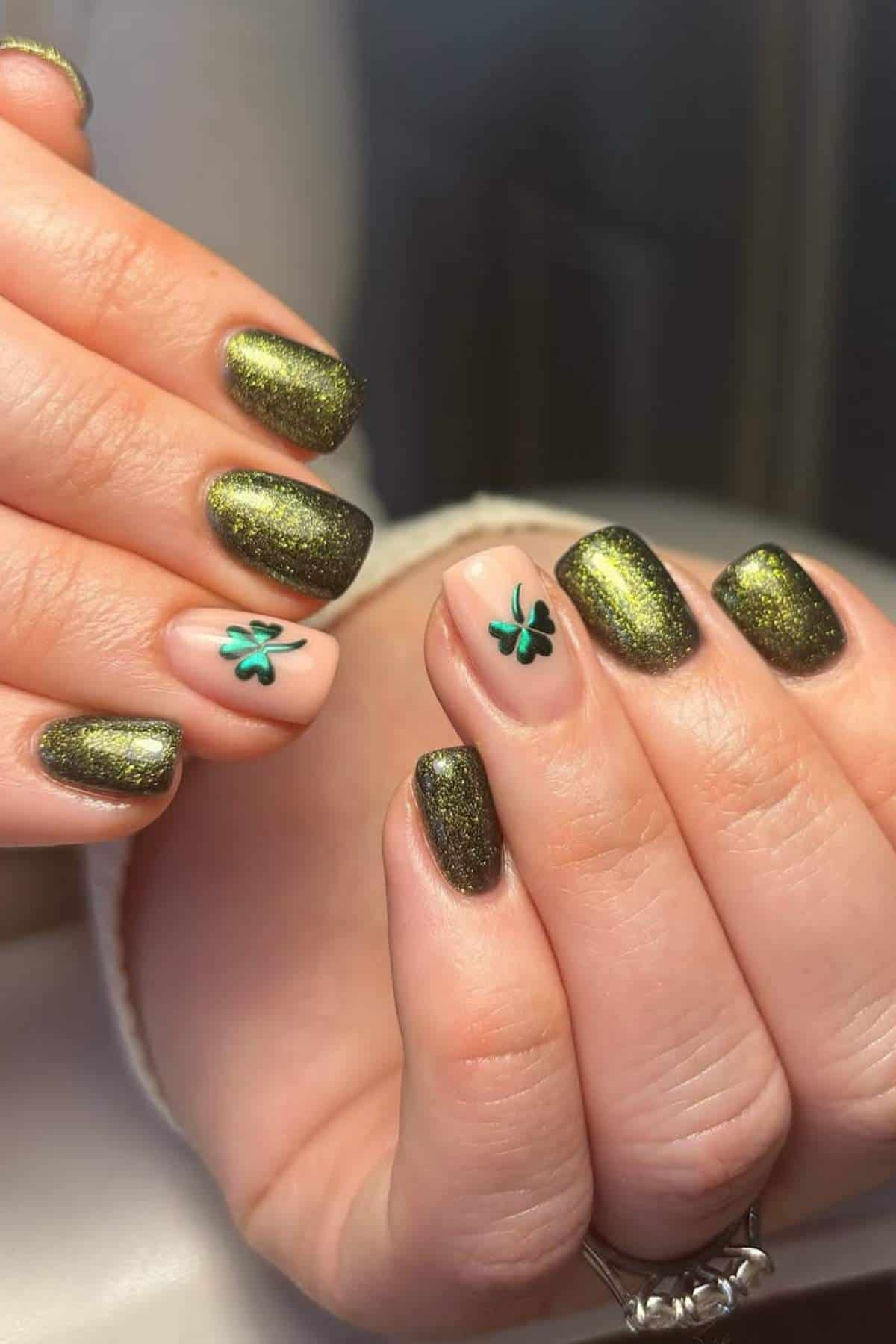 Hands with short, square-shaped nails painted metallic green, with two accent nails featuring small green shamrock designs on a nude base.