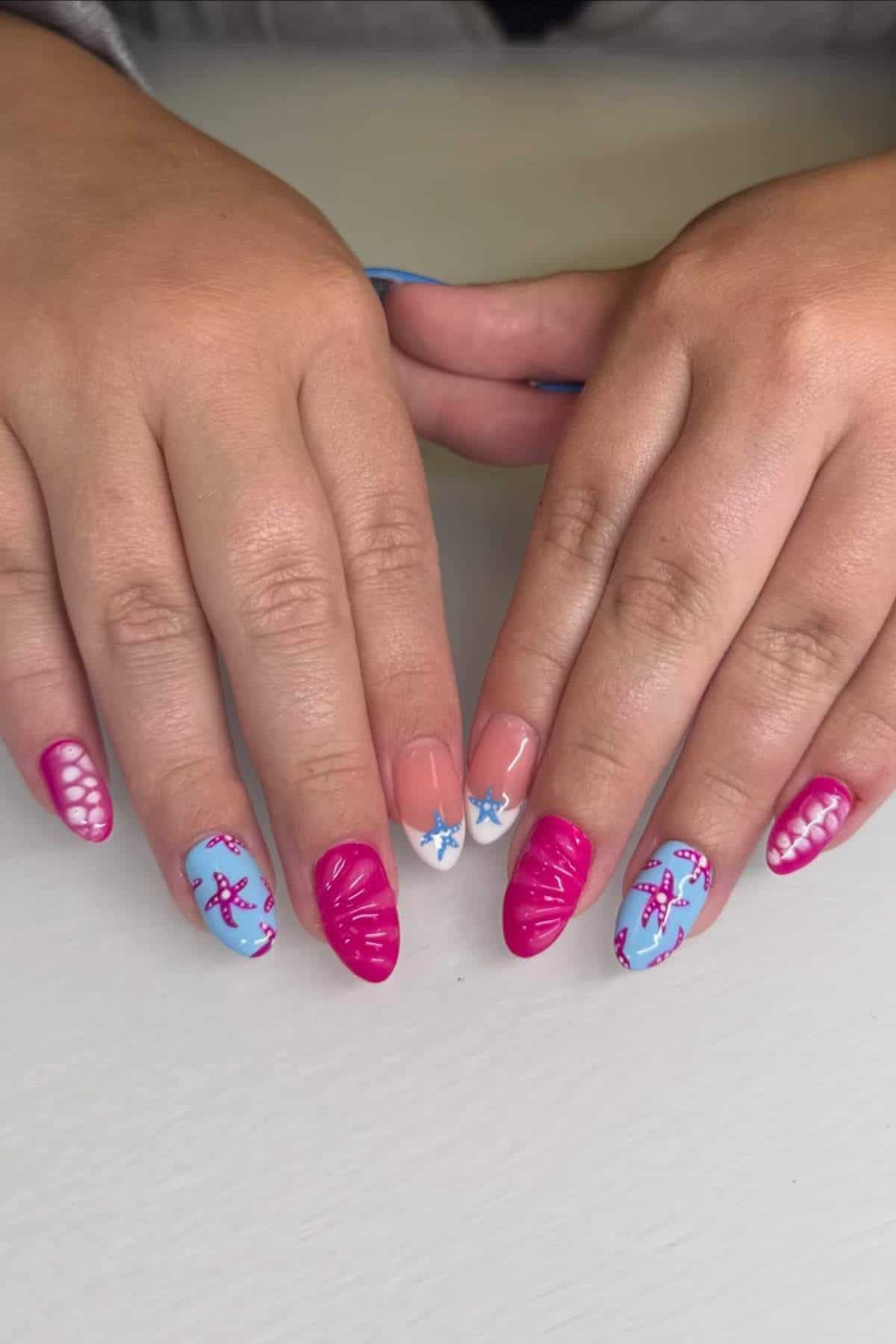 Hands with manicured nails featuring pink, blue, and white designs, including stars, swirls, and dotted patterns, displayed on a light surface.