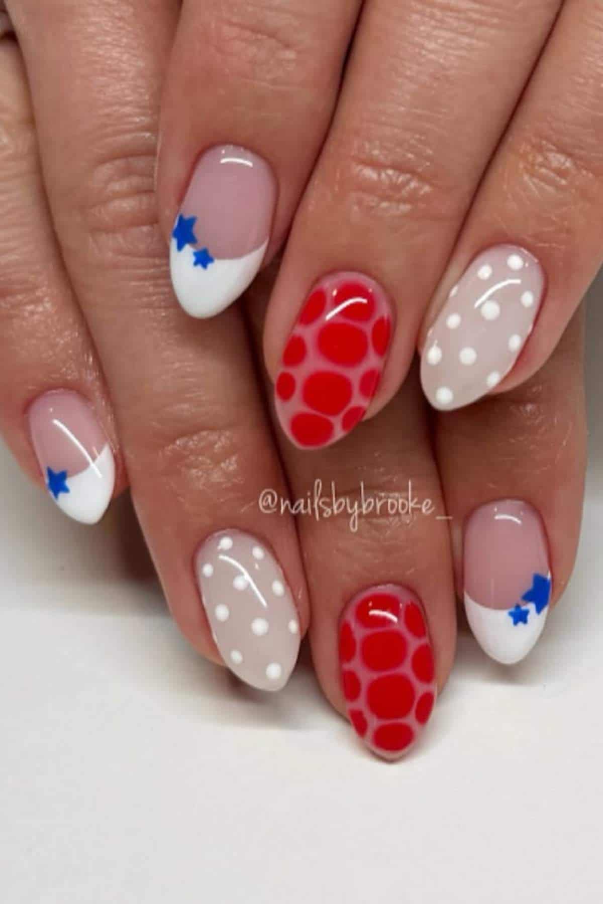 A hand with manicured nails featuring red polka dots, white tips with blue stars, and white polka dots on a pale base.