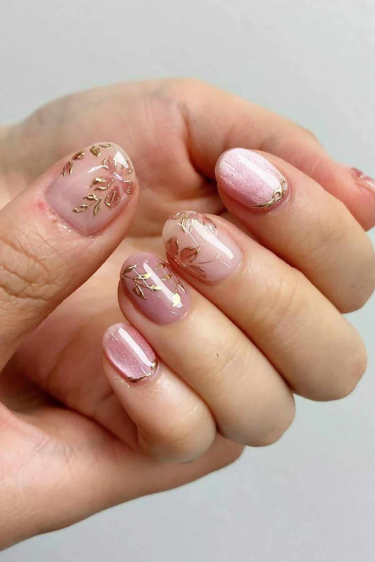 A hand with short, oval-shaped nails painted in pink polish and decorated with gold leaf patterns.