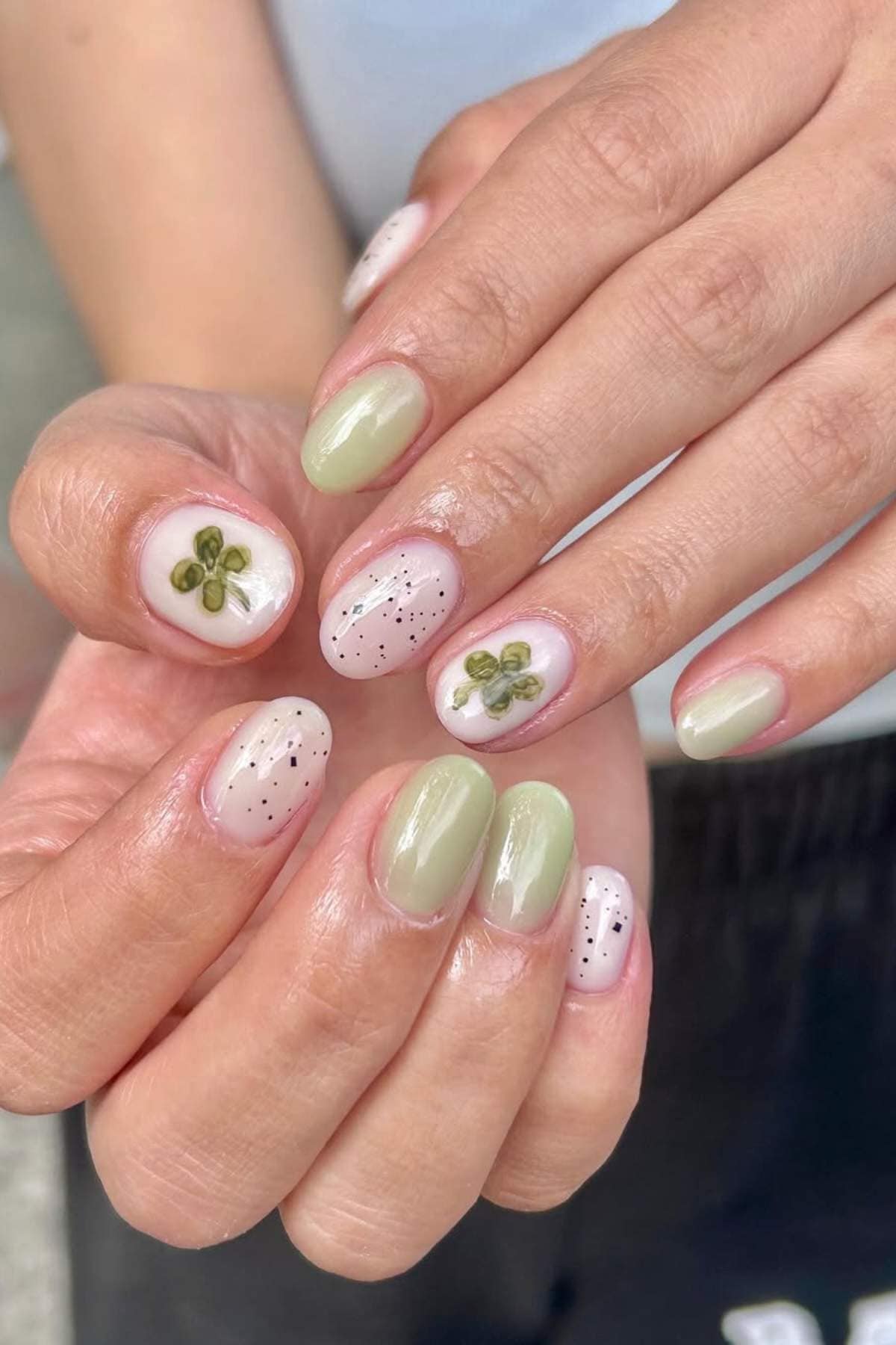 Hands with short nails painted in pale green and nude colors, featuring black dots and clover leaf designs on some nails.