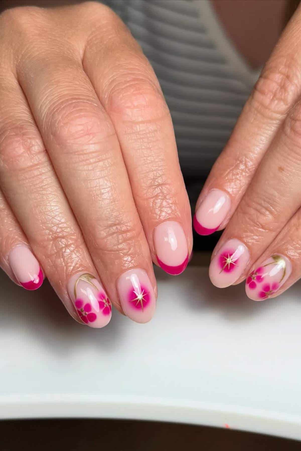 Hands with manicured nails featuring pink French tips and floral nail art with pink flowers and yellow centers on a light nude base.