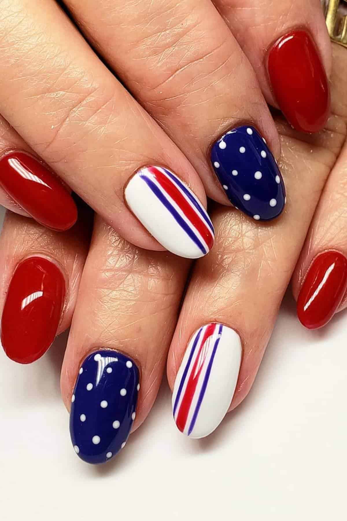 Hands with manicured nails featuring red polish, blue polish with white dots, and white polish with red and blue diagonal stripes.