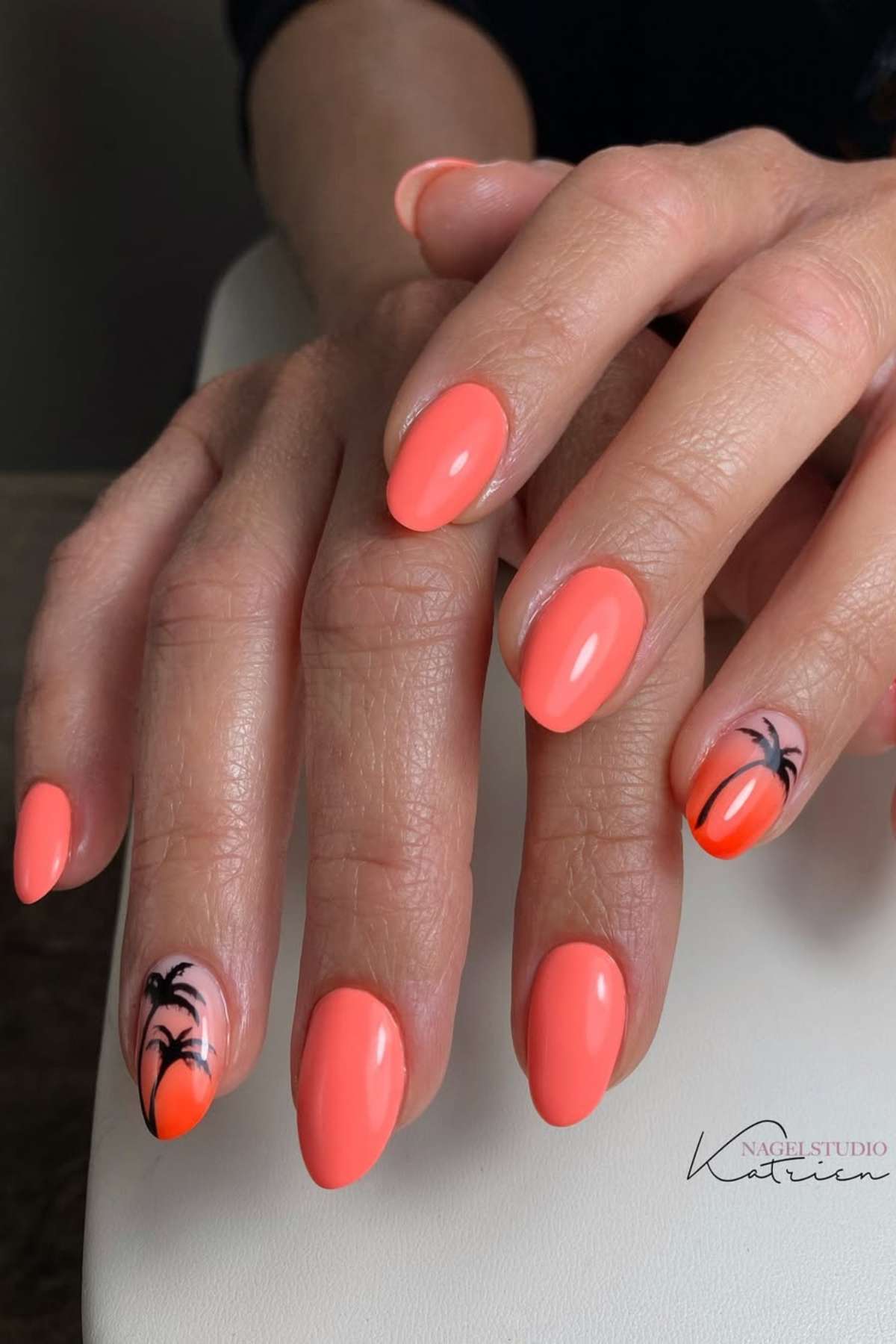 Hands with almond-shaped nails painted coral orange; ring fingernails feature black palm tree designs on a gradient orange background.