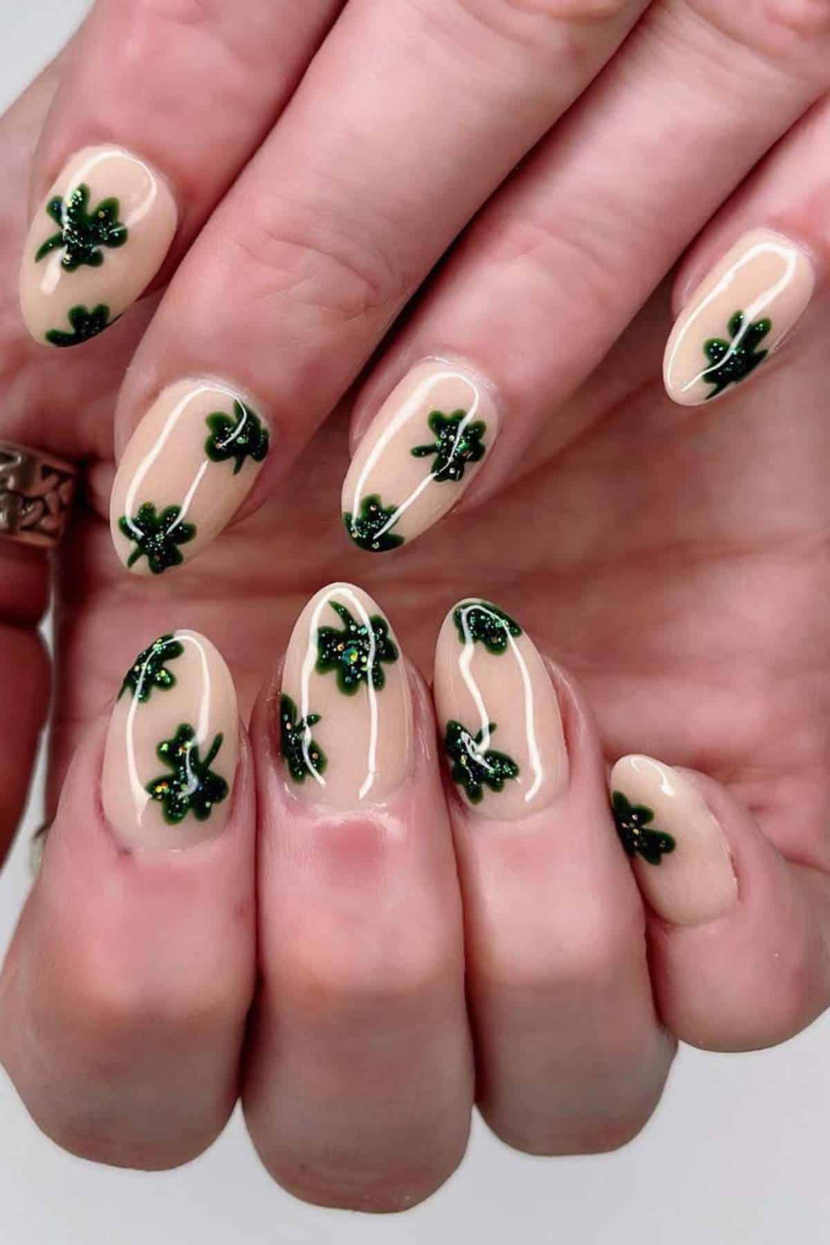 A hand with almond-shaped nails painted in a nude color, decorated with small green shamrock designs on each nail.