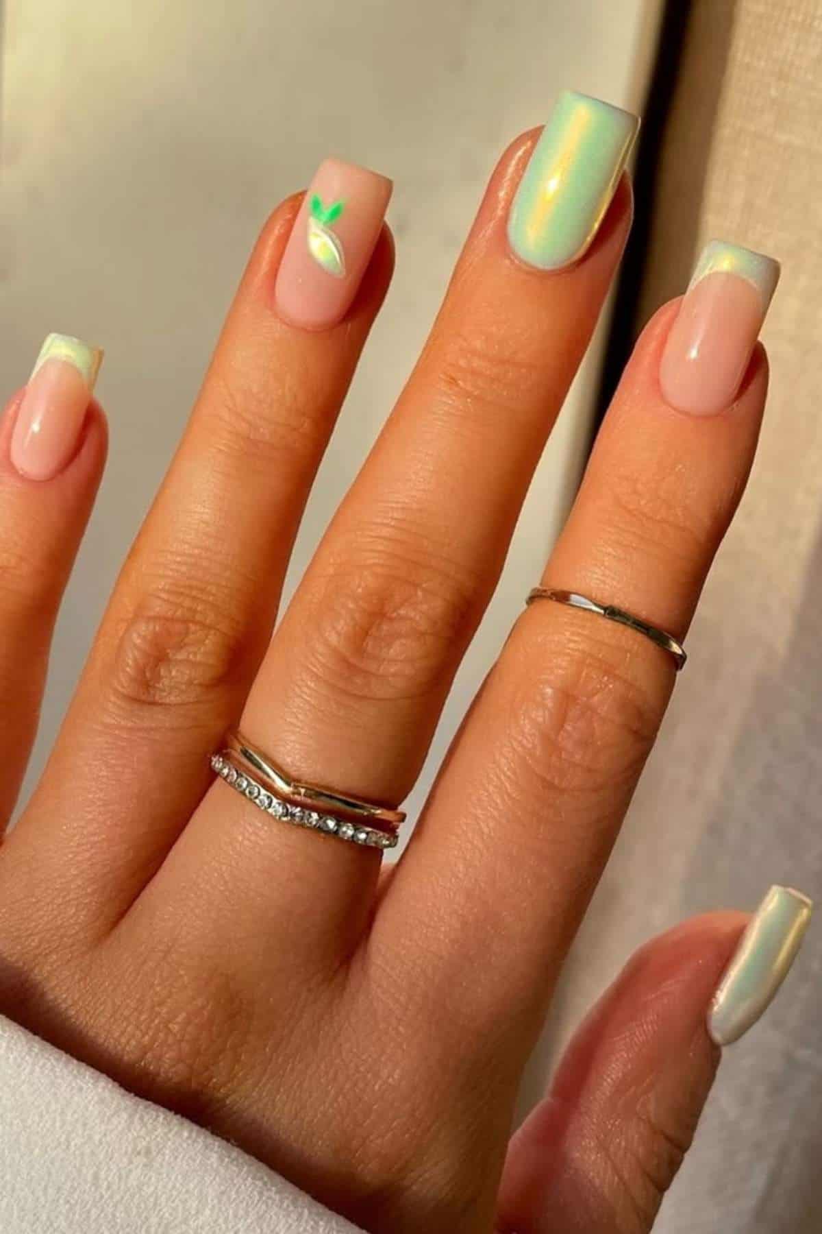 A hand with pale iridescent square-tipped nails, one featuring a small green leaf design, wearing three silver rings.