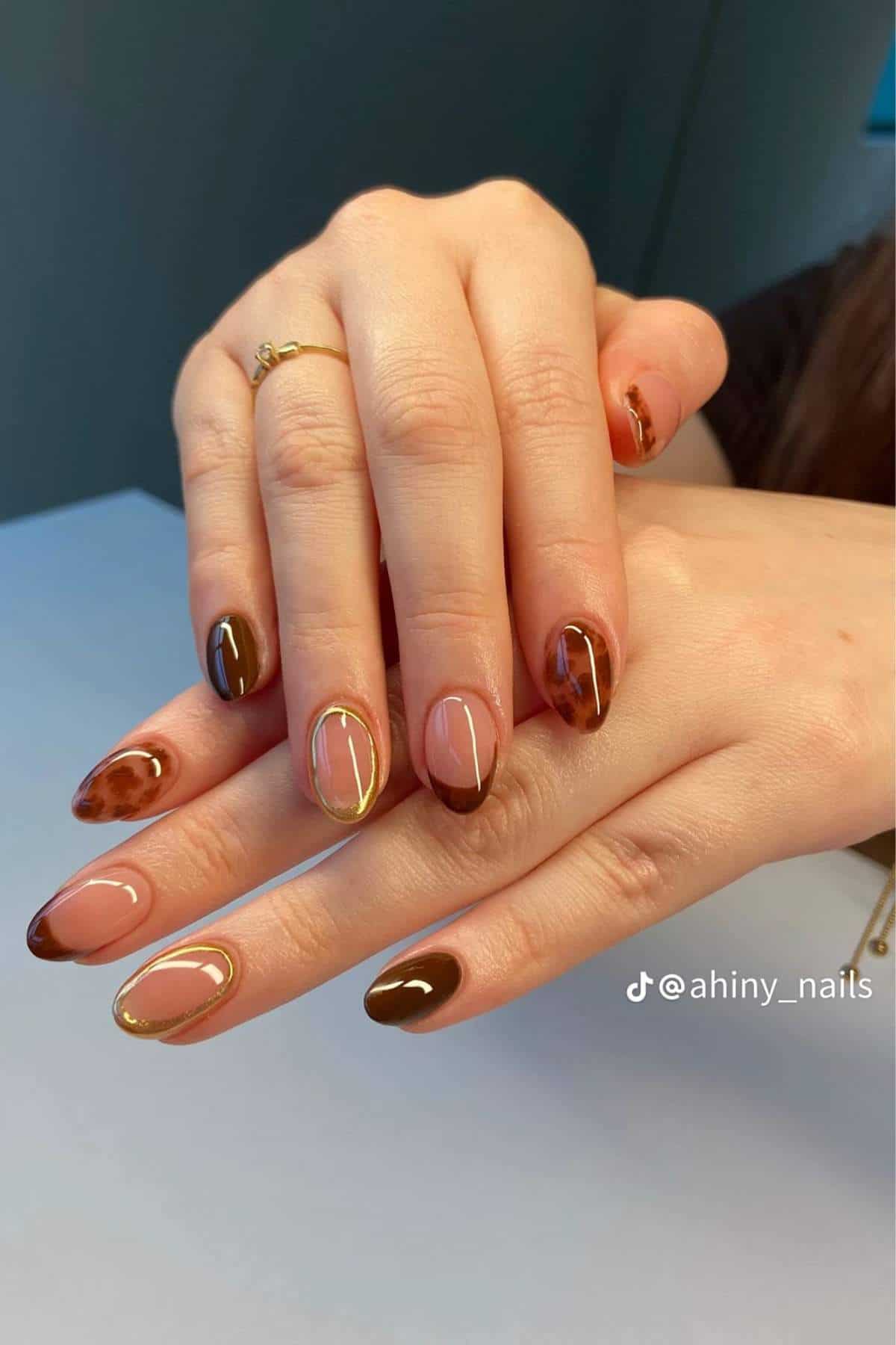 Hands with almond-shaped nails featuring a brown and nude abstract design, including gold accent lines and tortoiseshell patterns.