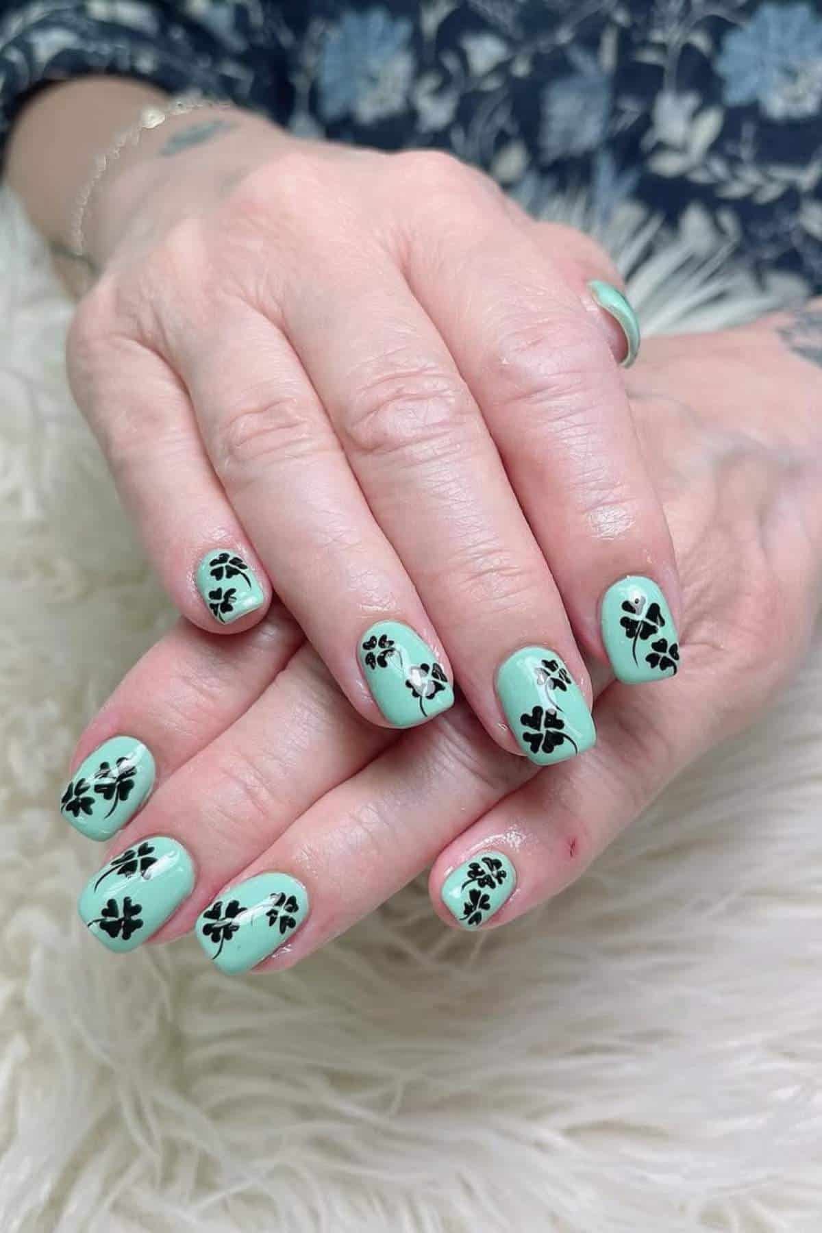 Hands with light green nail polish featuring black four-leaf clover designs on each nail, resting on a white textured surface.