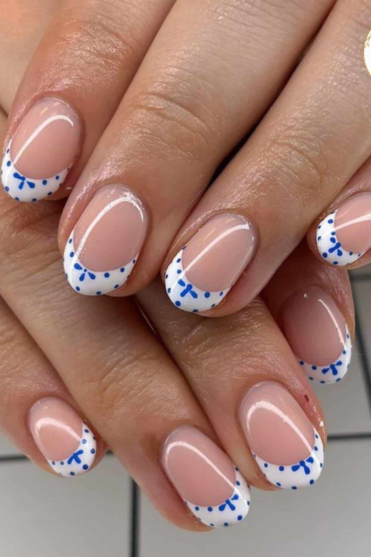 Close-up of hands with a French manicure featuring white tips and blue dotted and bow-like decorative accents on natural nails.