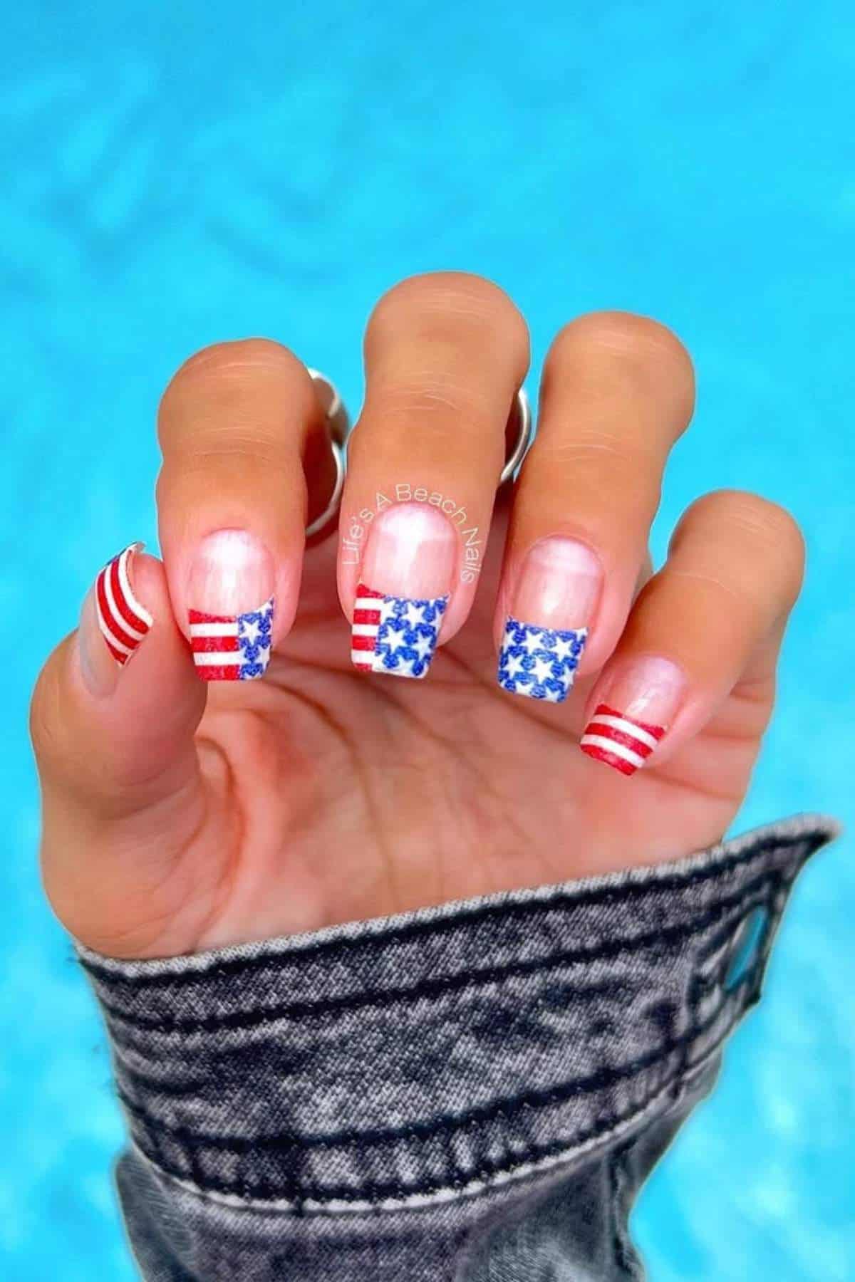 A hand with fingernails painted in an American flag design, featuring red and white stripes and blue with white stars, against a blue background.