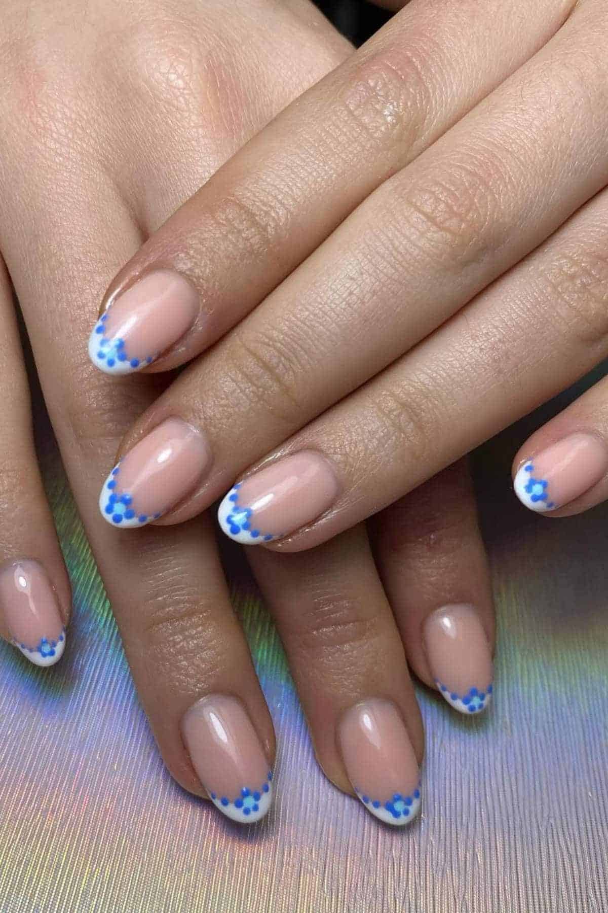 Hands with almond-shaped nails featuring a nude base and white French tips decorated with small blue flower designs.