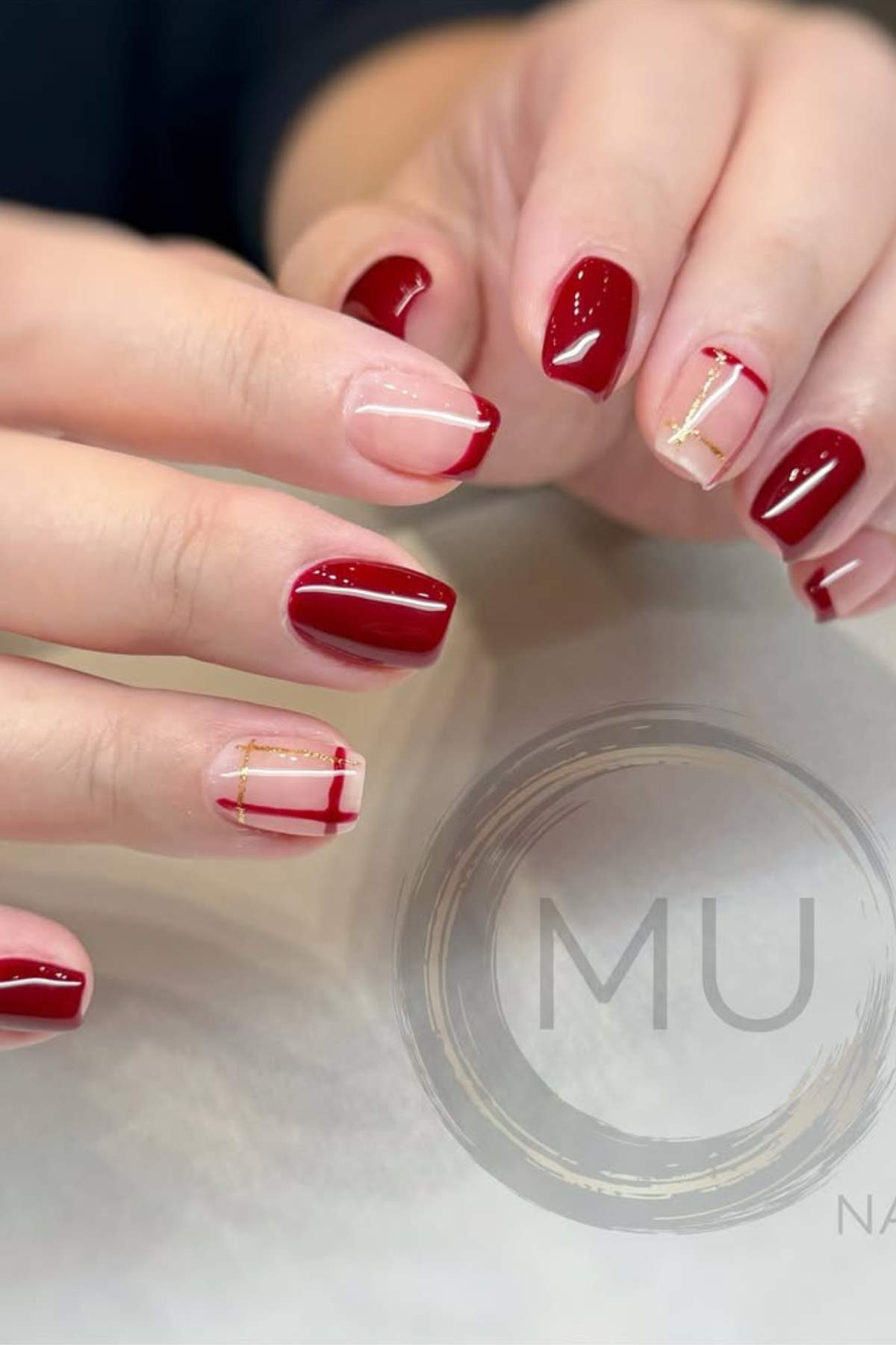 Hands with manicured nails featuring a mix of deep red polish and nude with red and gold geometric accents.