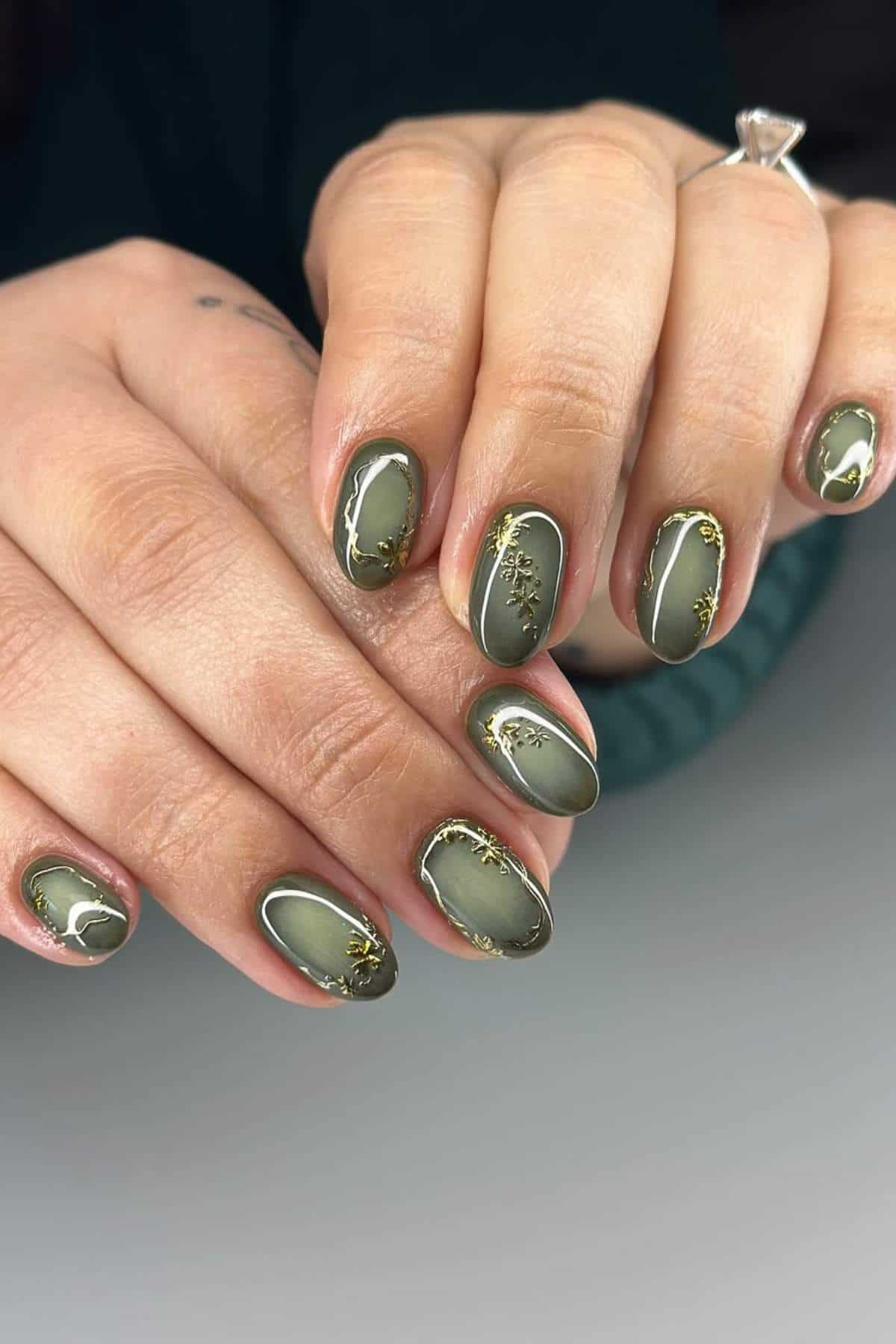 Close-up of hands with oval-shaped, olive green nails featuring gold foil accents and glossy finish.