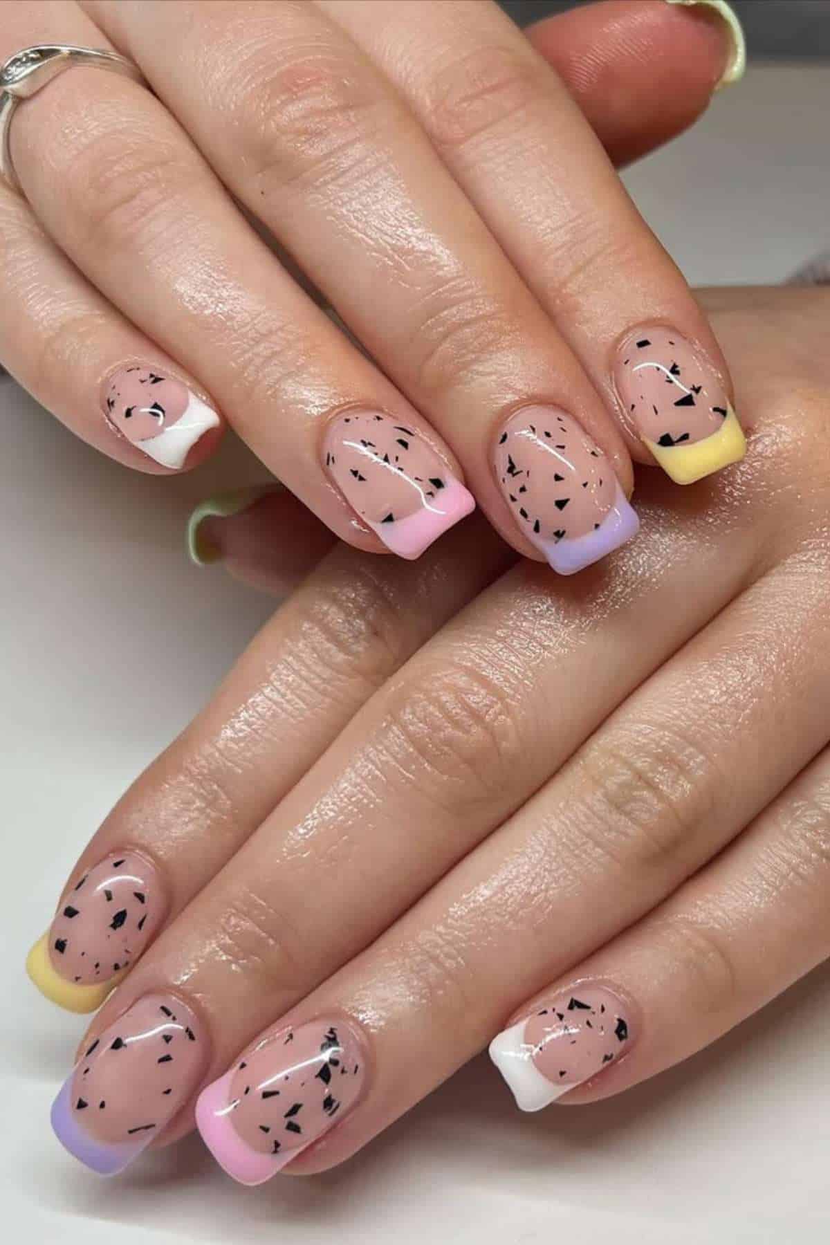 Hands with short square nails featuring pastel French tips, small black speckles, and a clear glossy base coat.