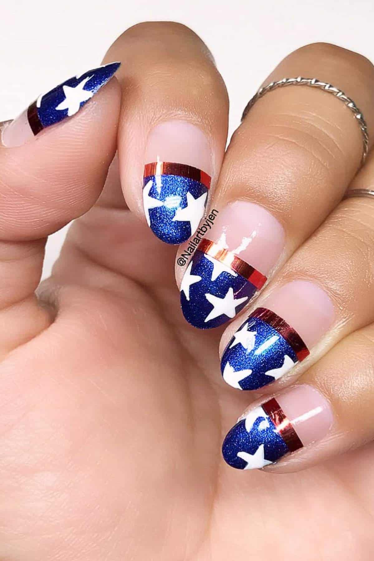 A hand with almond-shaped nails featuring blue glitter polish, white stars, and red metallic stripes designed in a patriotic theme.
