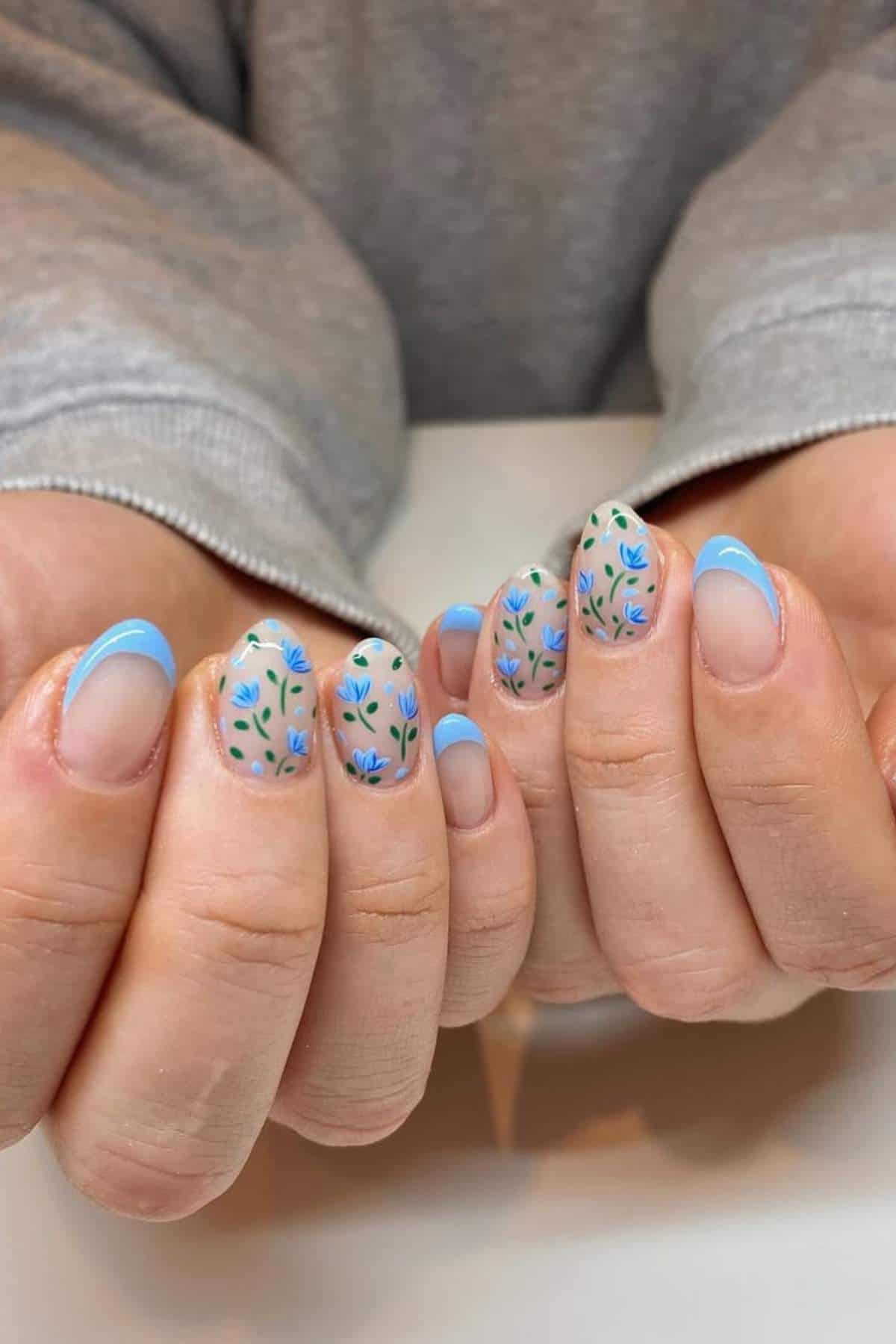 Hands with manicured nails featuring blue French tips and floral designs on a neutral base, displayed against a grey long-sleeve shirt.