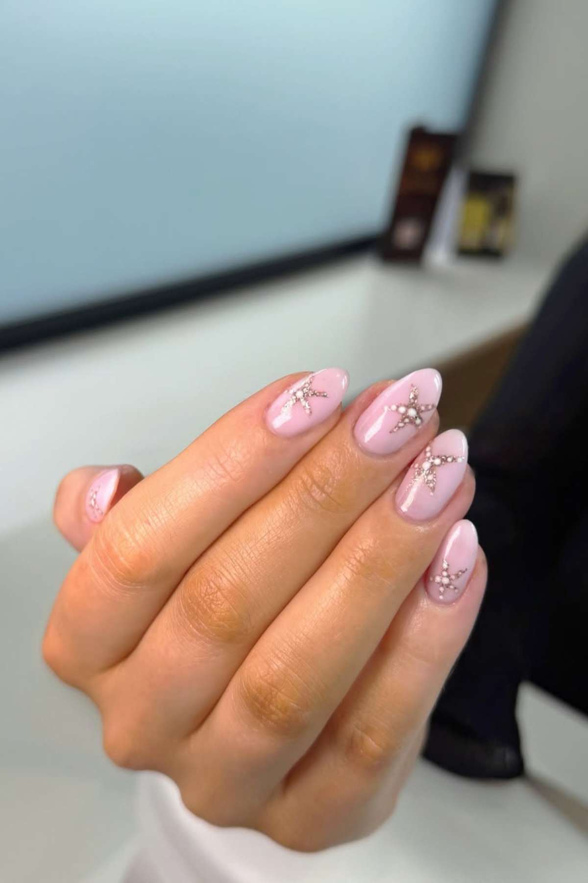 A hand with light pink almond-shaped nails featuring white line designs and small rhinestone accents, held up against a blurred indoor background.