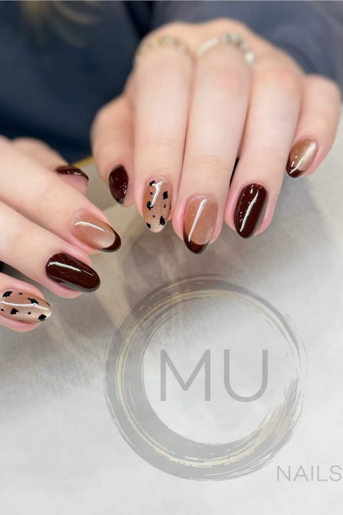 Hands with medium-length almond-shaped nails painted in brown and beige tones, featuring abstract black dot designs on two accent nails.