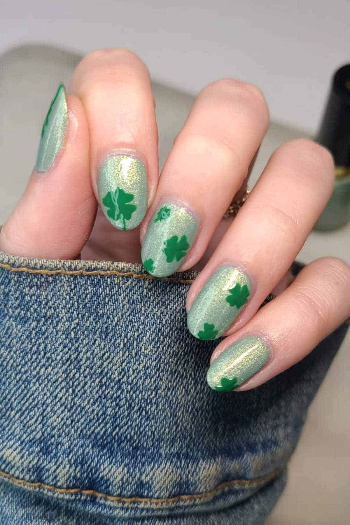 A hand with light green, shimmery nail polish featuring green shamrock designs on each nail, resting on denim fabric.