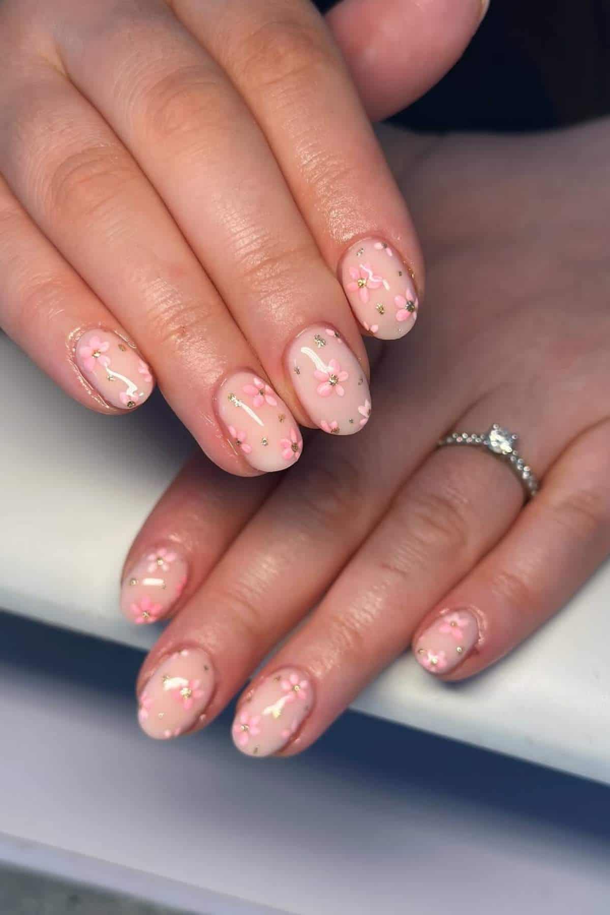 Hands with a silver ring on one finger, featuring short, almond-shaped nails with pink and white floral nail art on a nude base.