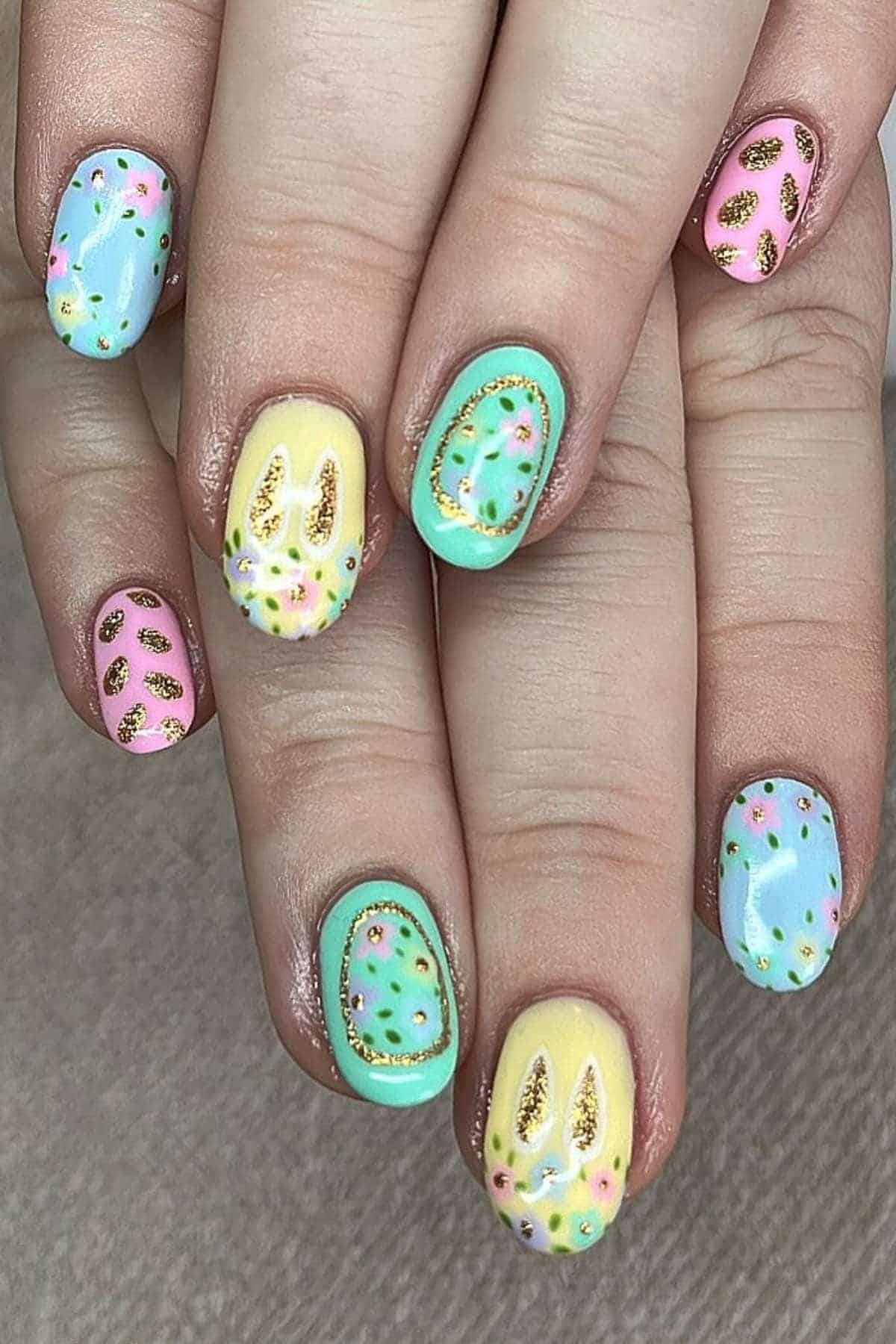 Hands with pastel-colored nail art featuring bunny ear designs, floral accents, and gold details on each fingernail.
