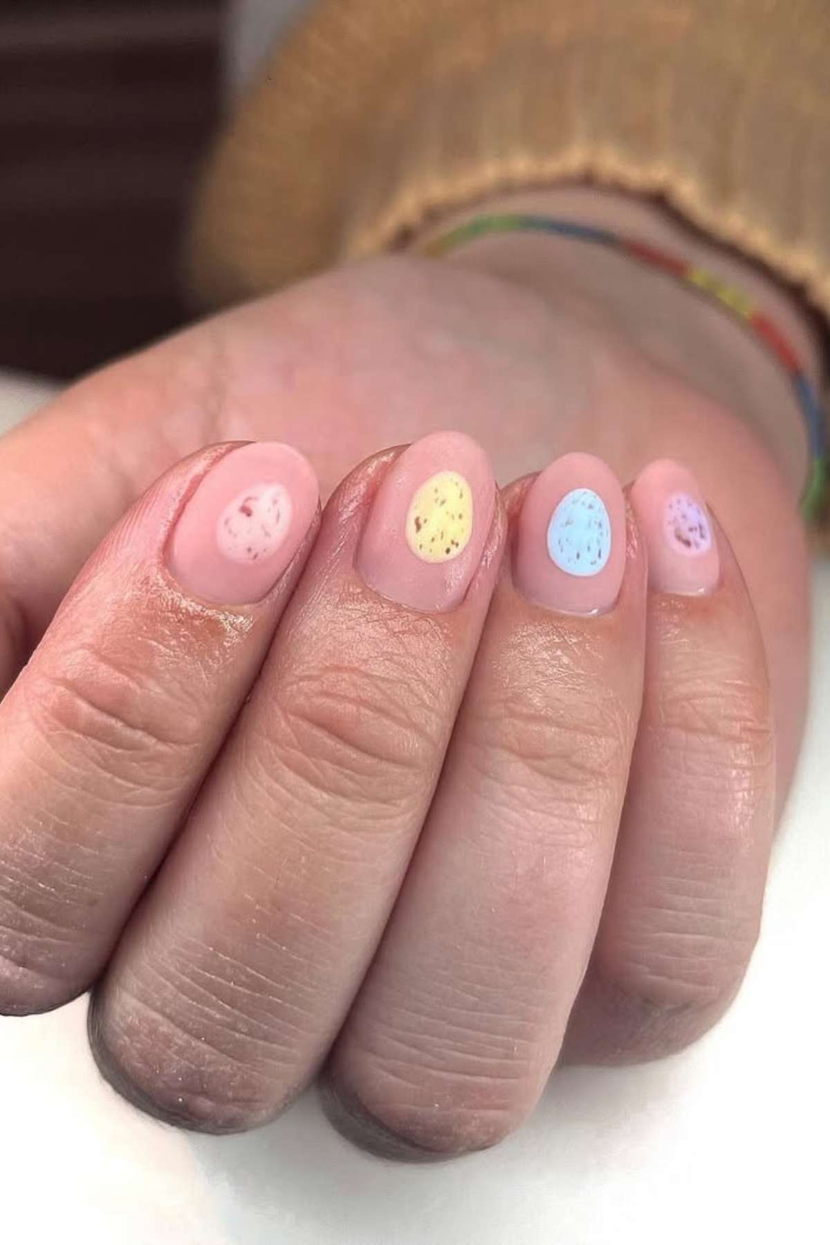 A hand with short, oval-shaped nails painted light pink, each featuring a small pastel oval with black specks resembling Easter eggs.