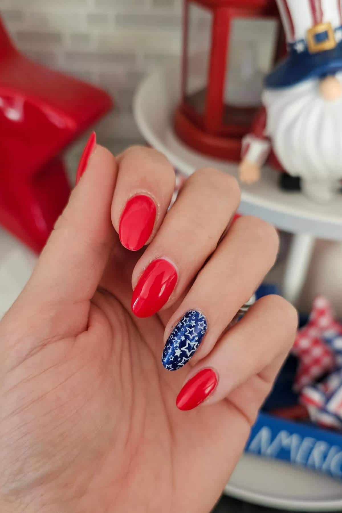 A hand with red-painted fingernails; the ring fingernail features blue polish with a white star pattern. Patriotic-themed decor is blurred in the background.