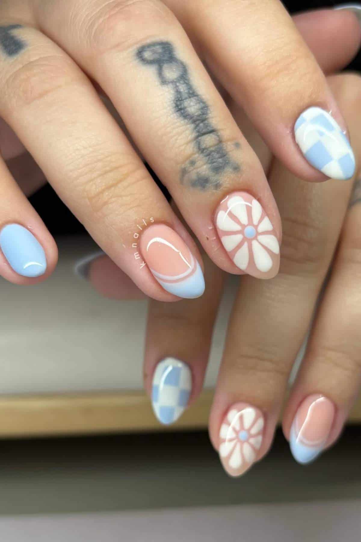 Hands with almond-shaped nails painted in pastel blue, white, and nude tones featuring checkered, floral, and abstract designs. A tattoo is visible on one of the fingers.