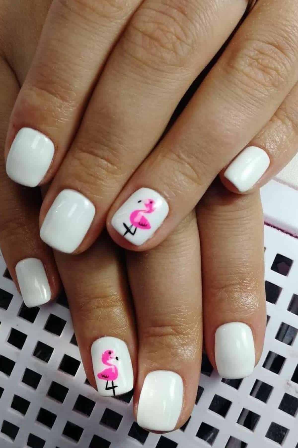 Hands with short, square-shaped white painted nails; the ring fingers feature a small pink flamingo design on each nail.