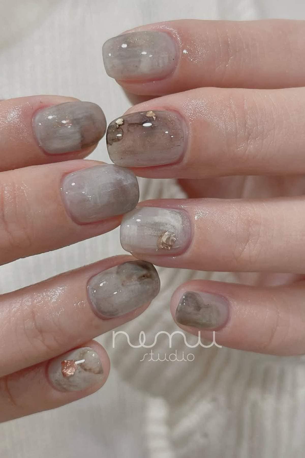Close-up of hands with short, rounded nails featuring a gray and brown marble design with glossy finish and subtle metallic foil accents.