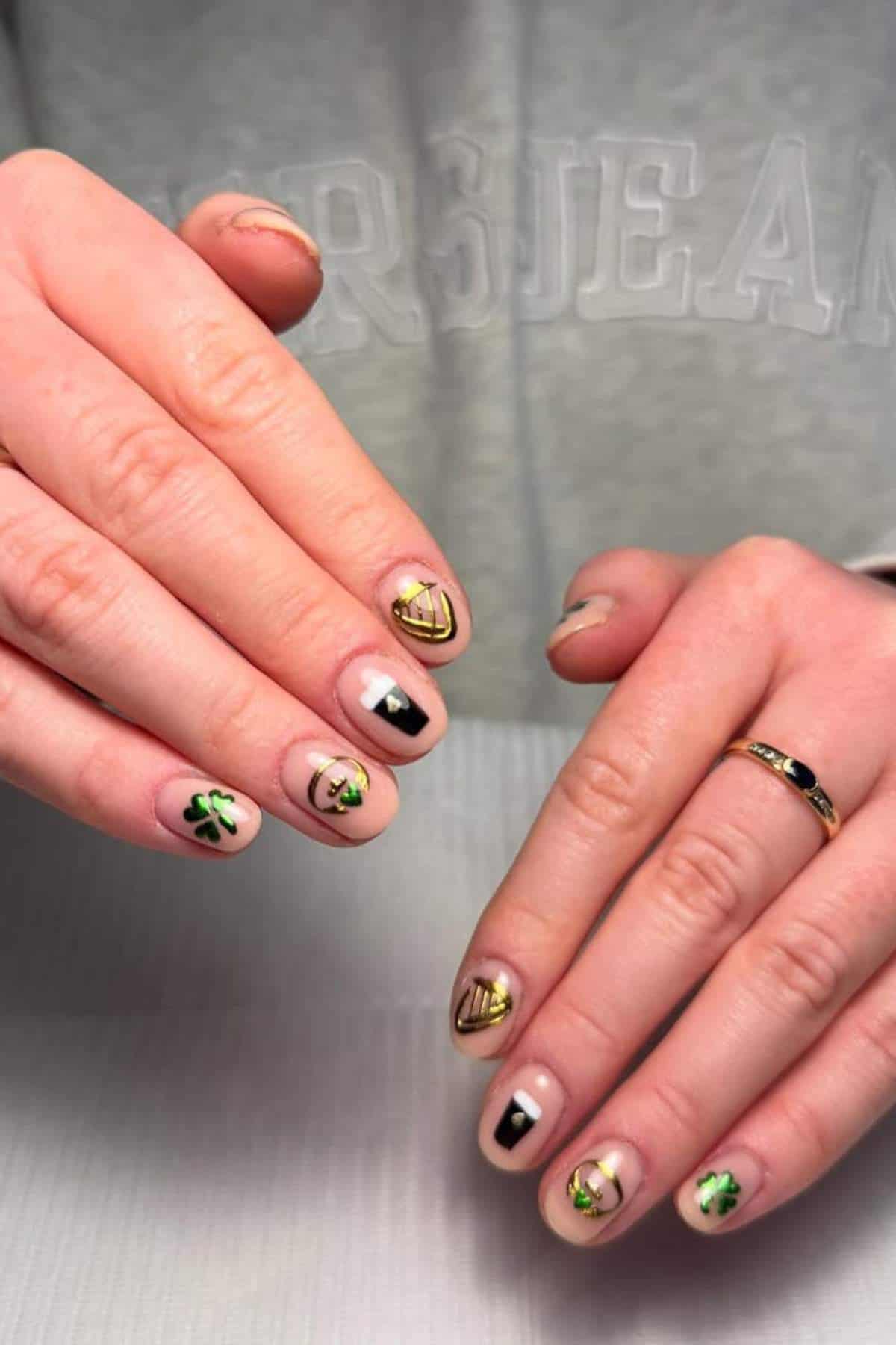 Two hands with short nails, each featuring different nail art designs in black, gold, and green on a nude base. The person is wearing a light sweatshirt with visible text.