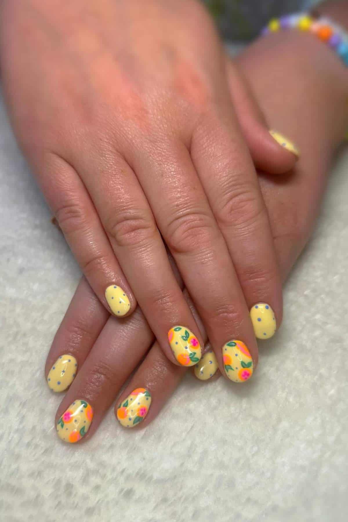 Hands with yellow nail polish; some nails feature colorful floral and fruit designs, while others have small polka dots. Nails are short and rounded.