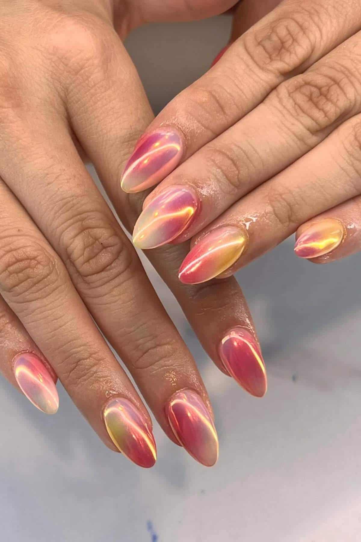 Hands with almond-shaped nails painted in a shiny, iridescent pink and gold gradient design, with a glossy finish.