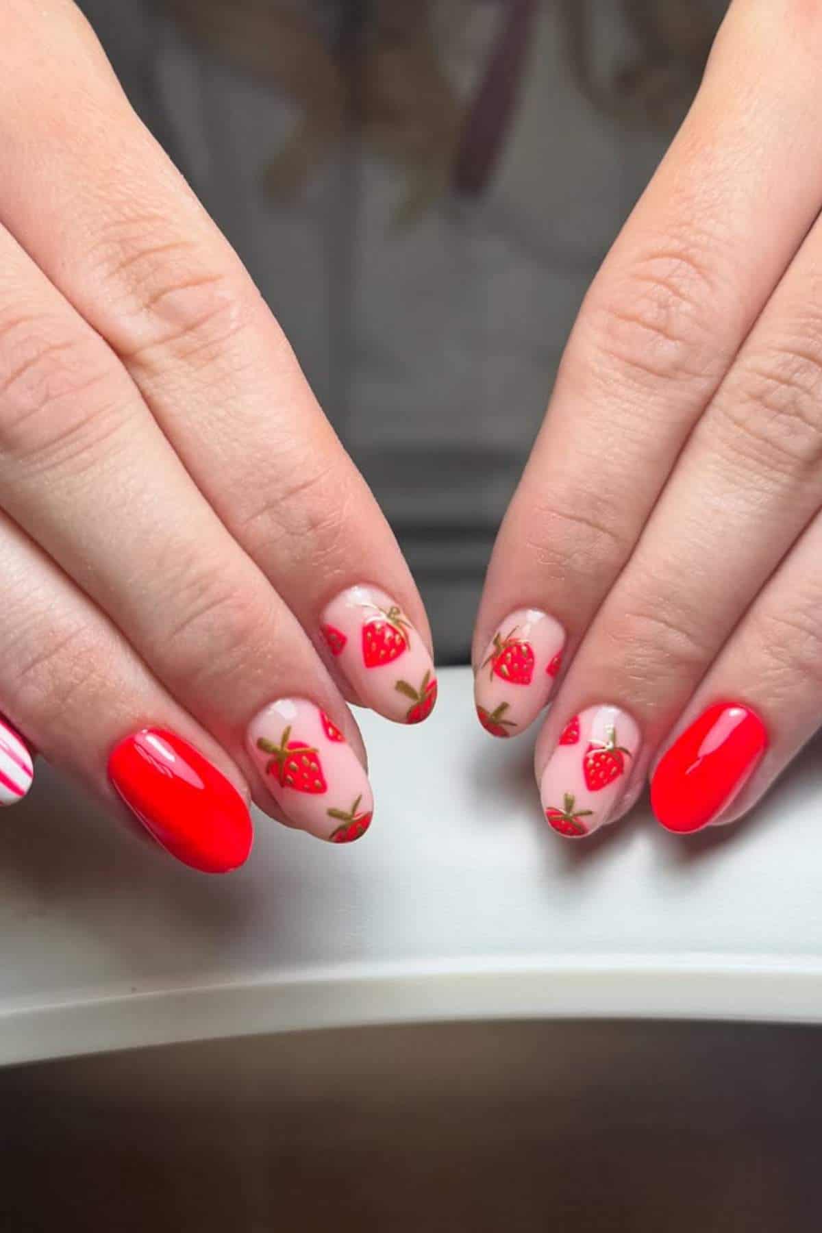 Hands with manicured nails featuring red polish, strawberry designs on a nude base, and a red-and-white striped accent nail.