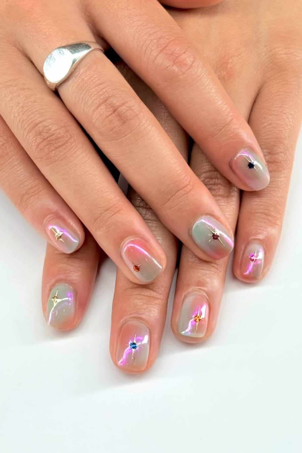 Hands with short, oval-shaped nails painted with a sheer nude polish, featuring multicolored starburst designs and small gem accents on each nail. Silver ring on one finger.